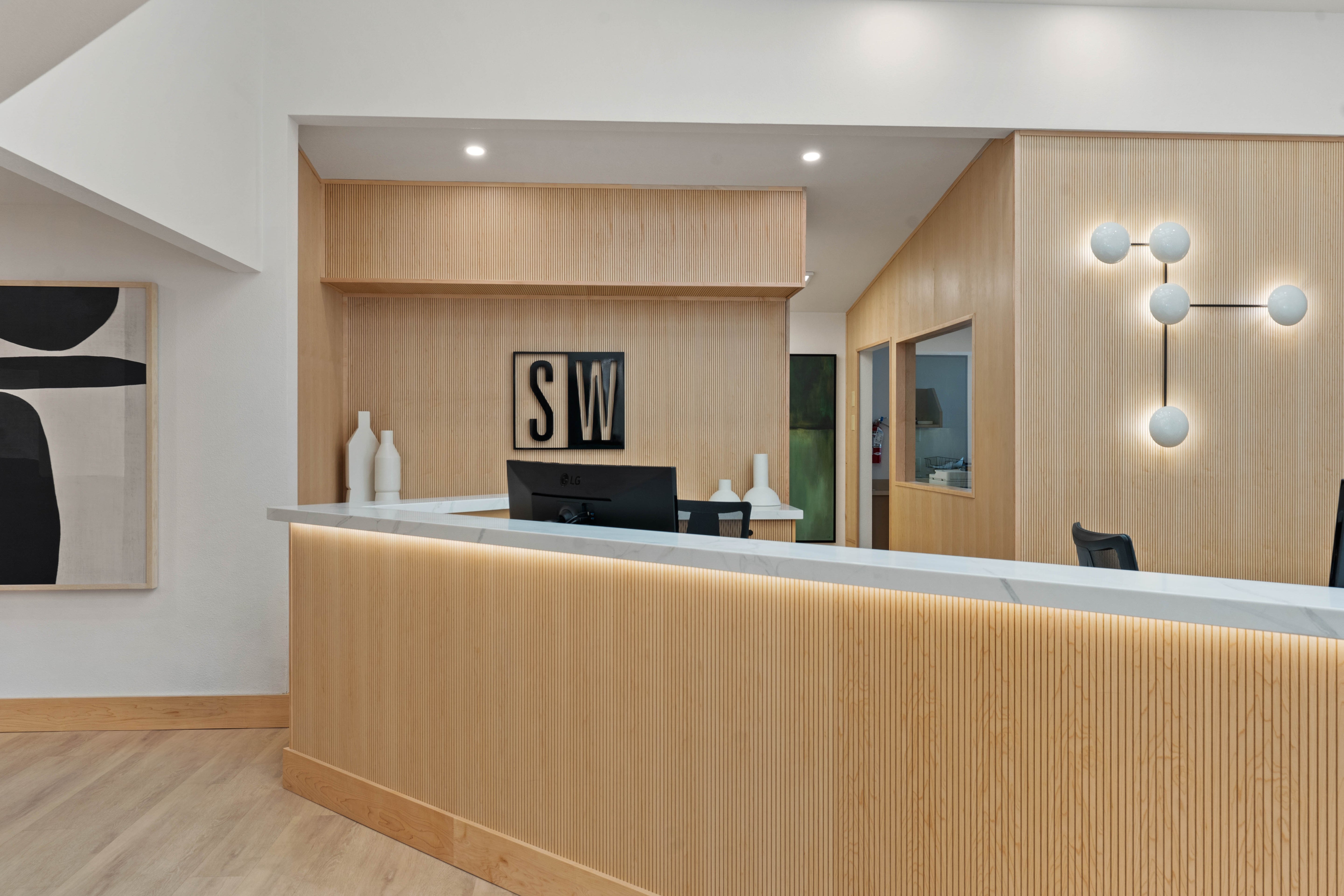 Bright reception area with fluted light wood accents, black brand logo on back wall, white marble countertops, geometric light fixtures, natural skylight illumination.