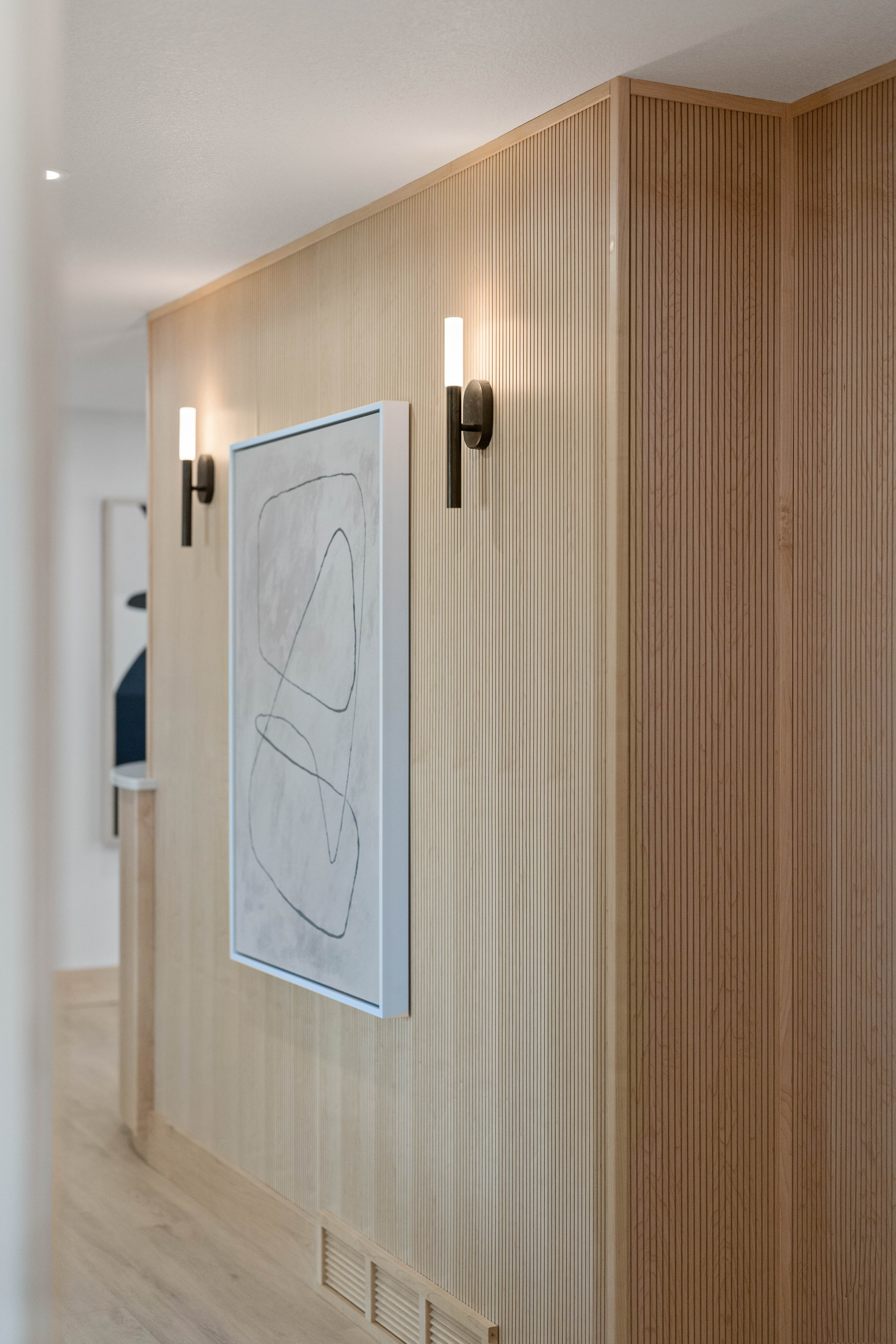 Close-up of modern hallway with minimalist wall art, fluted light wood paneling, and sleek wall sconces.