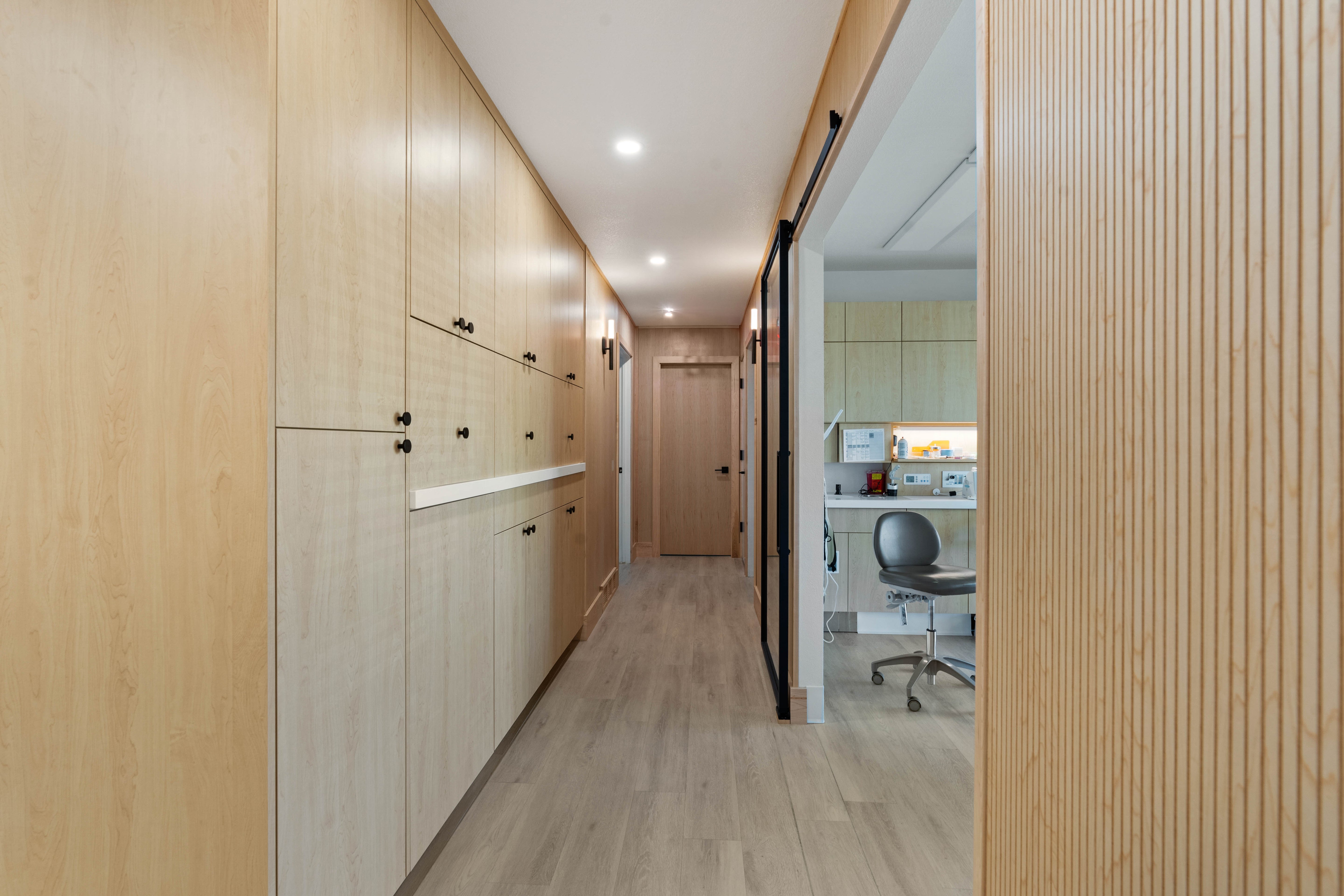 Hallway with built-in wood cabinetry, fluted light wood walls, sliding glass doors leading to a private room, and modern light fixtures.