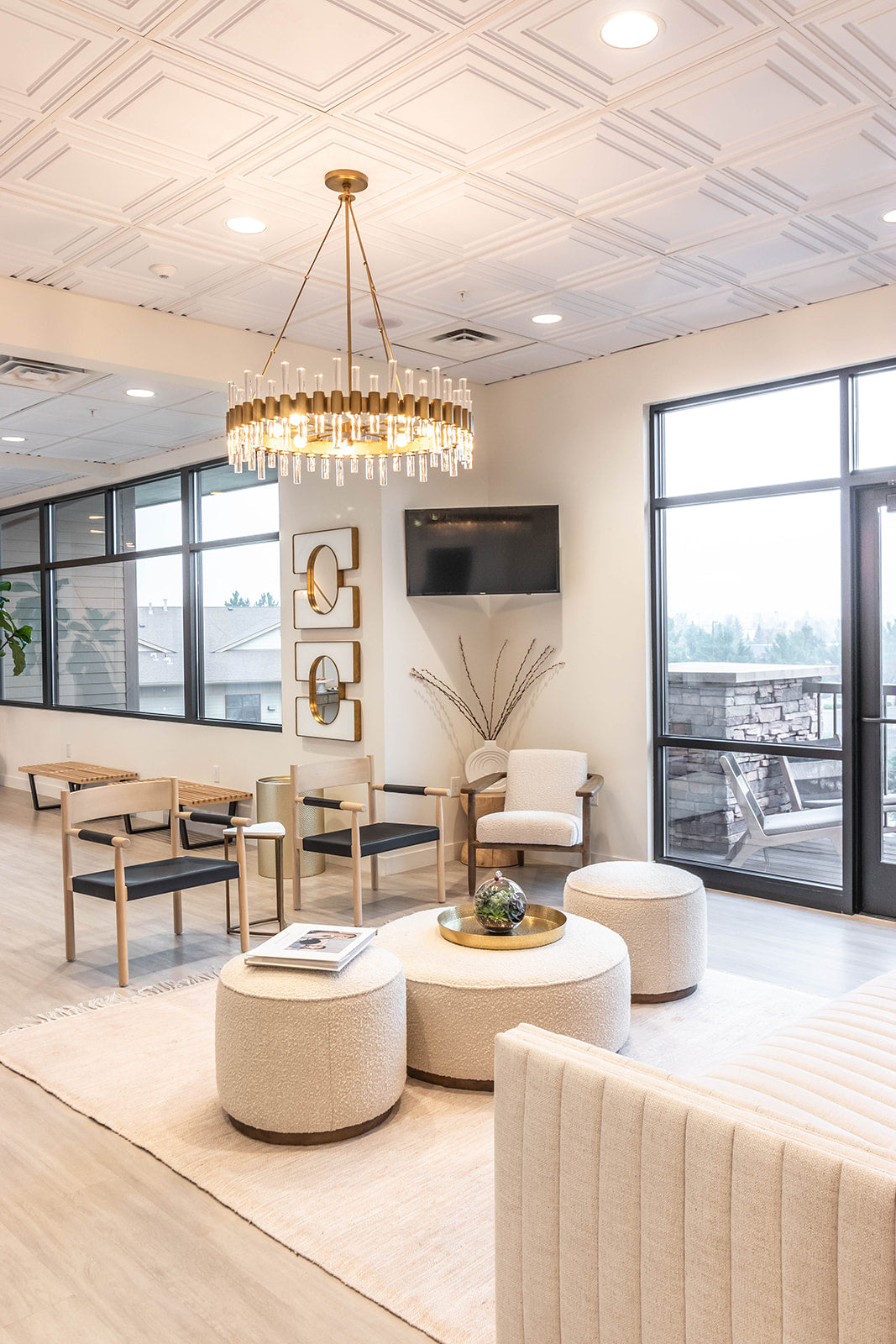 Bright waiting area with plush seating, a gold chandelier, neutral furniture and decor with gold accents, and large windows overlooking an outdoor terrace.