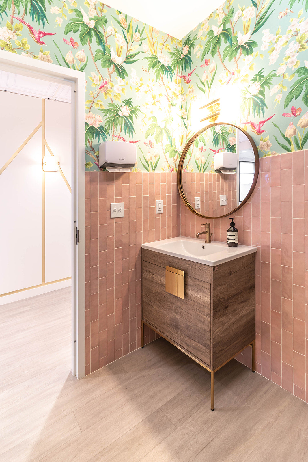 Vibrant restroom with floral wallpaper, pink tiles, and a wood vanity with gold hardware, creating a refreshing and stylish space.