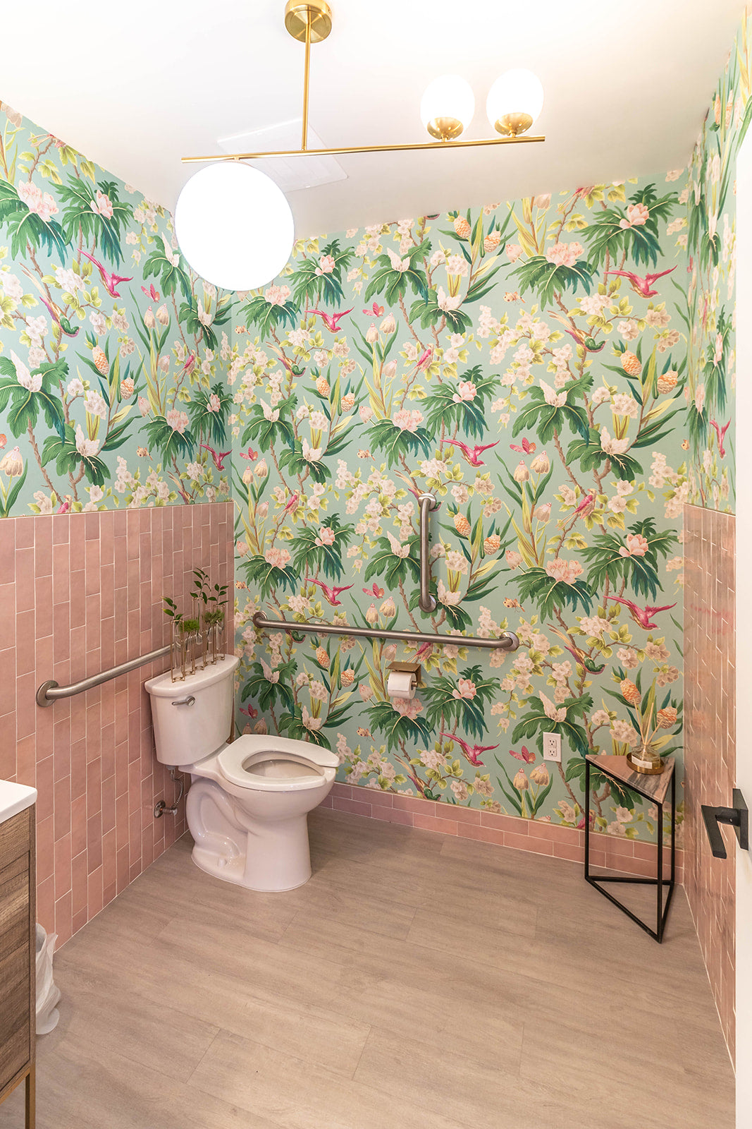 Vibrant restroom with floral wallpaper and pink subway tiles, featuring gold light fixtures and accessibility railings.