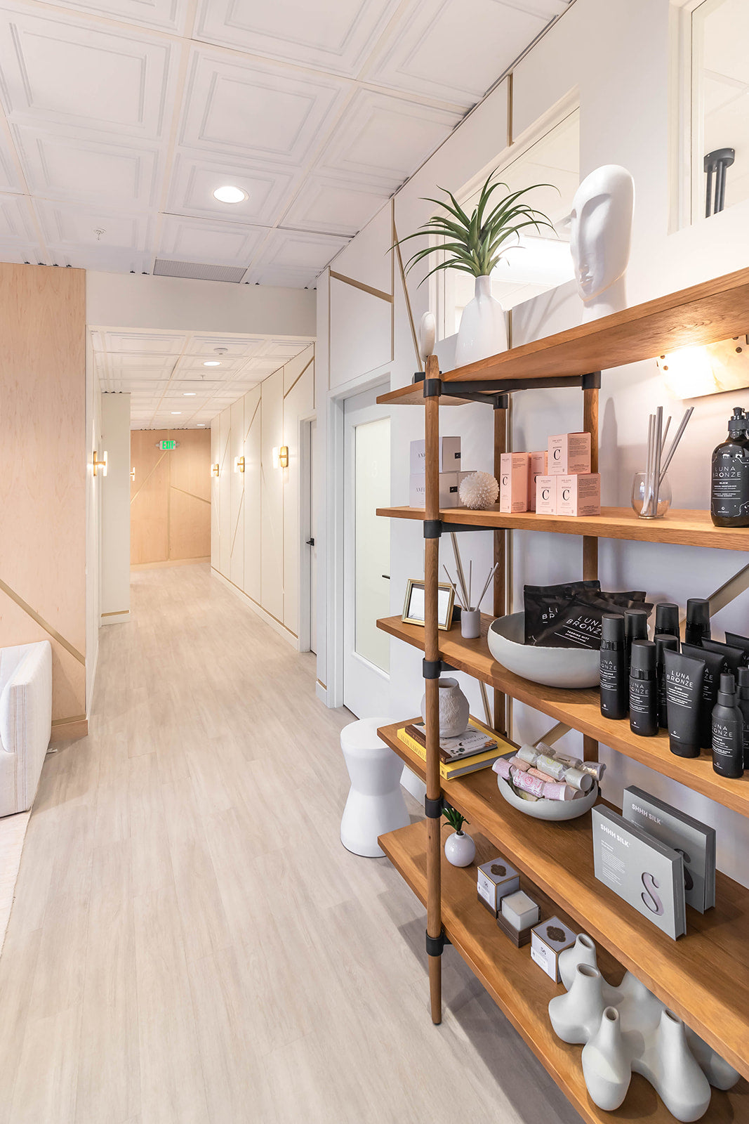 Elegant hallway with white walls featuring gold lines leading to private treatment rooms, and open wooden shelving displaying premium skincare products.