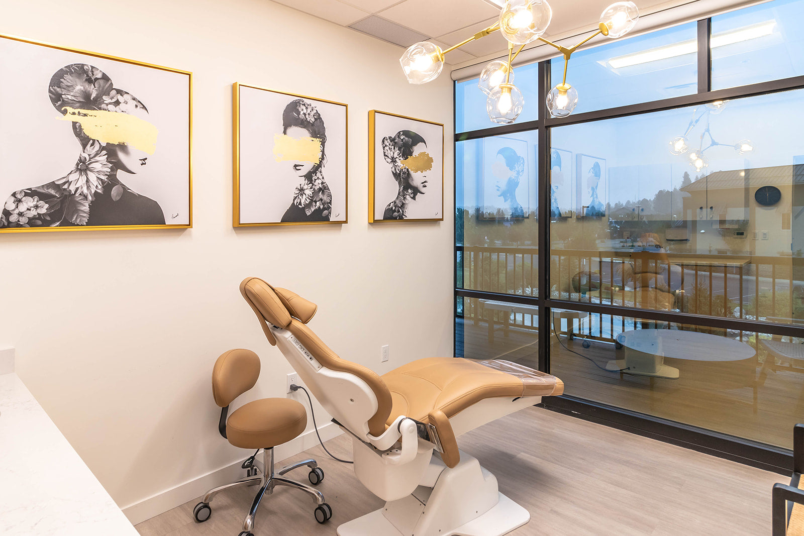 Bright treatment room, black and white framed art with gold accents on wall, a comfortable tan leather recliner chair, and large windows offering a view of the outdoor balcony.