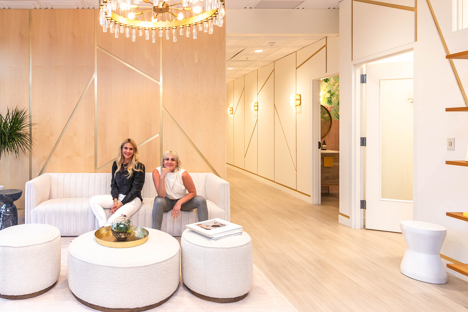 Two women seated on a white sofa in a stylish and well-lit reception area with geometric wall designs, gold accents, and a chandelier overhead.