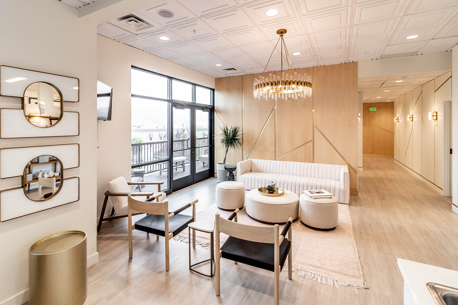 Elegant waiting area featuring plush seating, striking chandelier, neutral furniture and decor with gold accents, and access to an outdoor balcony through large glass doors.