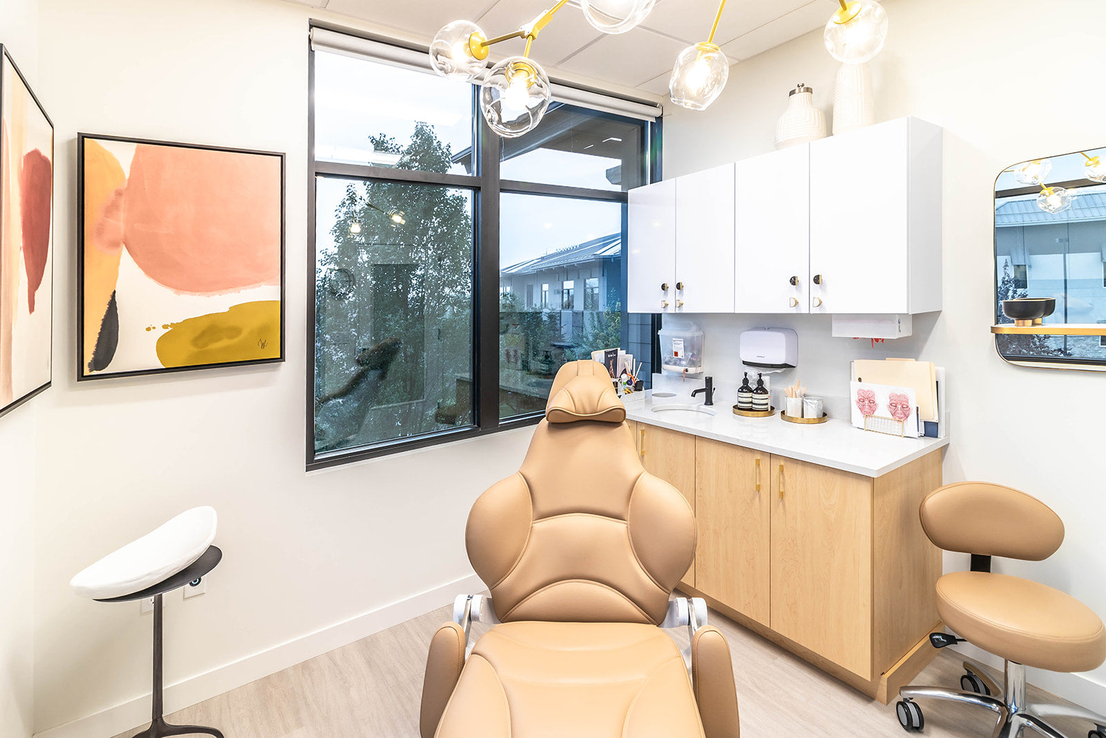 Bright treatment room with a tan reclining chair, abstract wall art, gold light fixture, and large windows offering natural light.