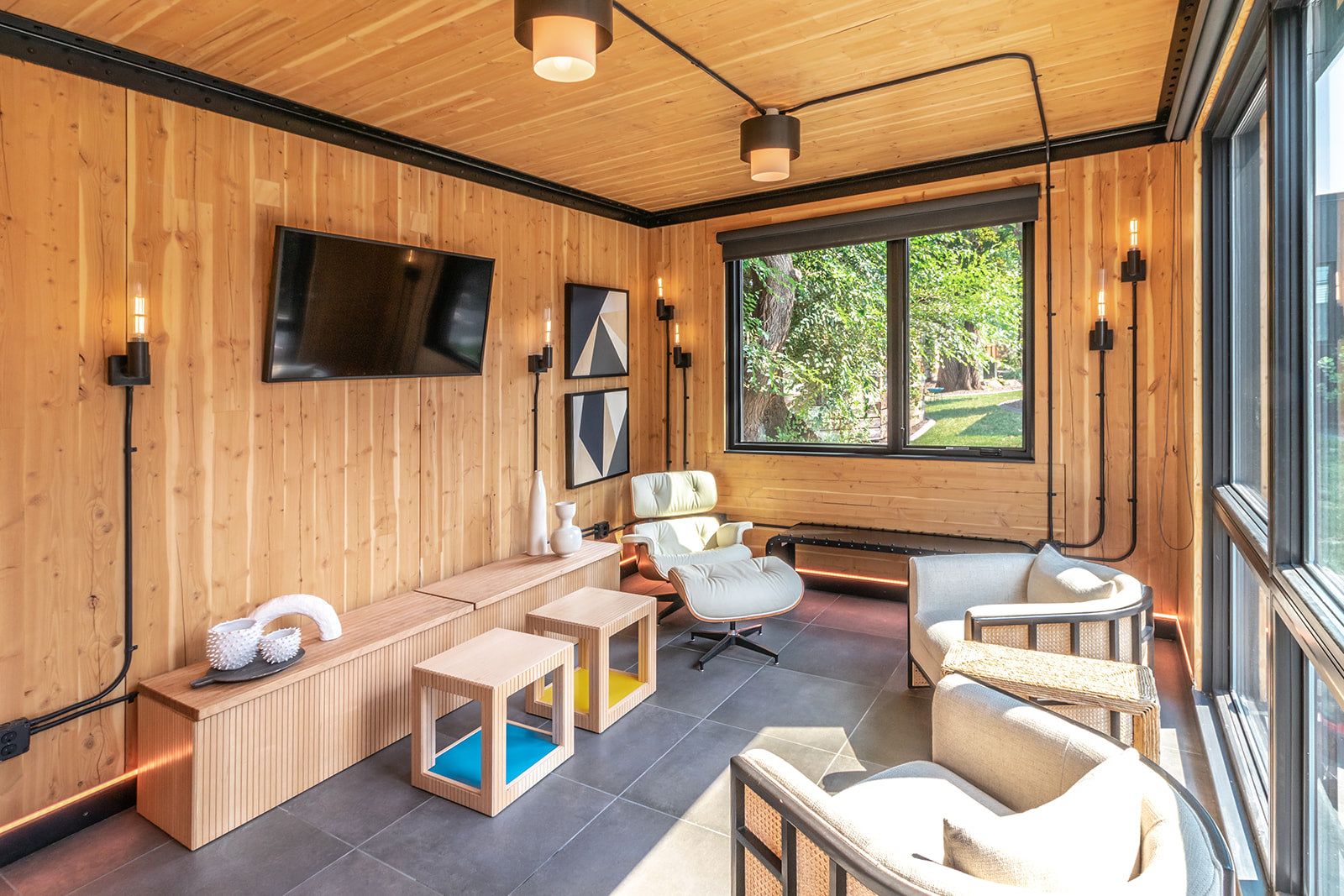 Cozy living room with wood-paneled walls, neutral-color seating, a wall-mounted TV, and large windows offering natural light.