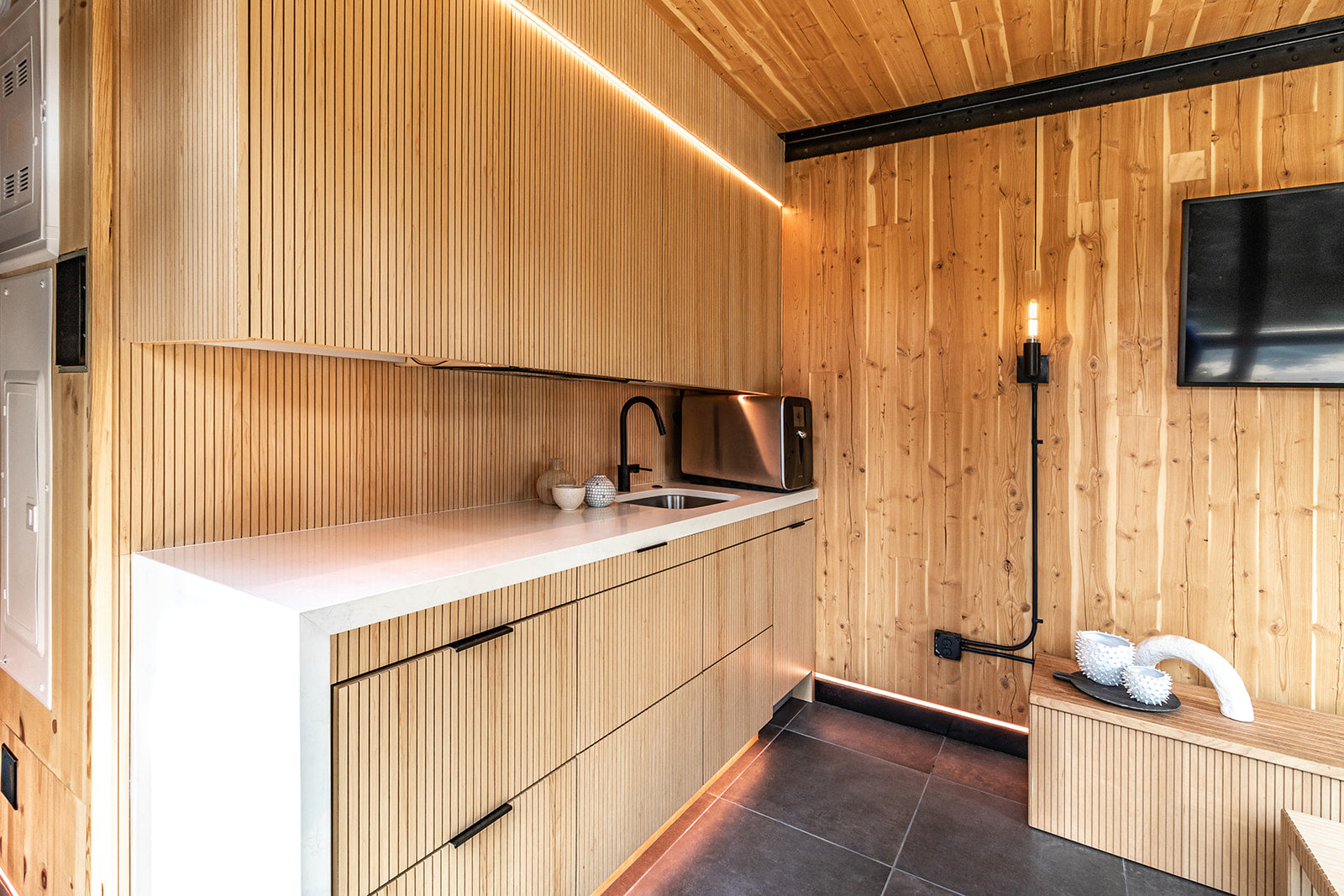 Kitchenette with light wood paneling on cabinets, black fixtures, under-cabinet lighting, light wood walls all around.