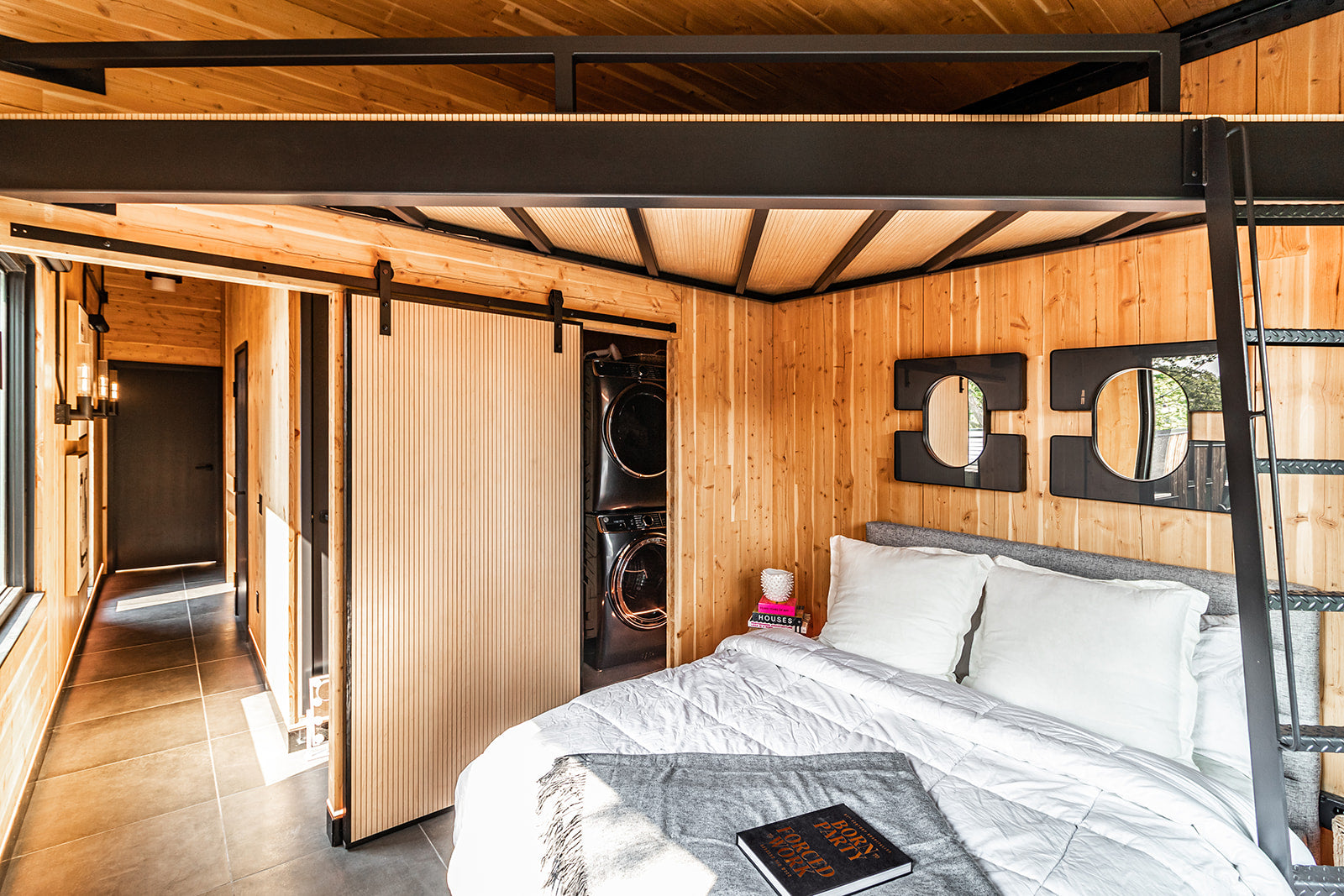 Bedroom with wood paneled walls all around, metal platform above bed with black metal stairs, two mirrors above headboard, and sliding fluted wood door leading to laundry room.