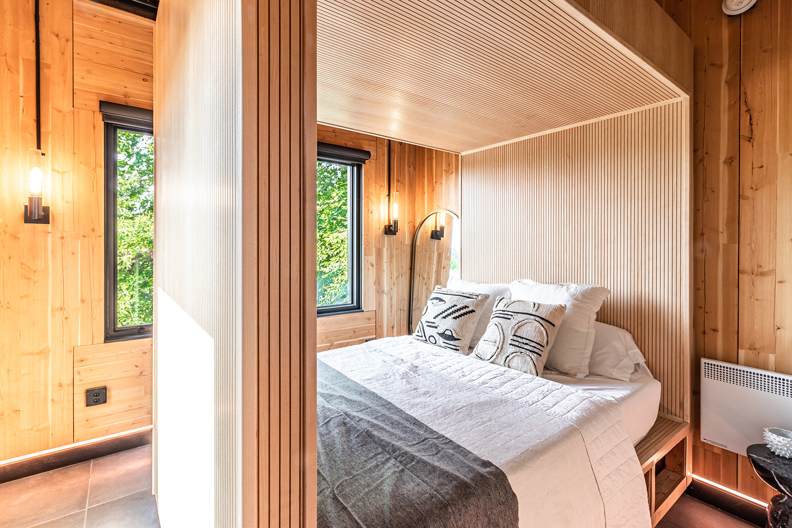 Bedroom with fluted light wood structure going around the bed, wood paneled walls all around, white and gray bedding, and large window looking out into nature.