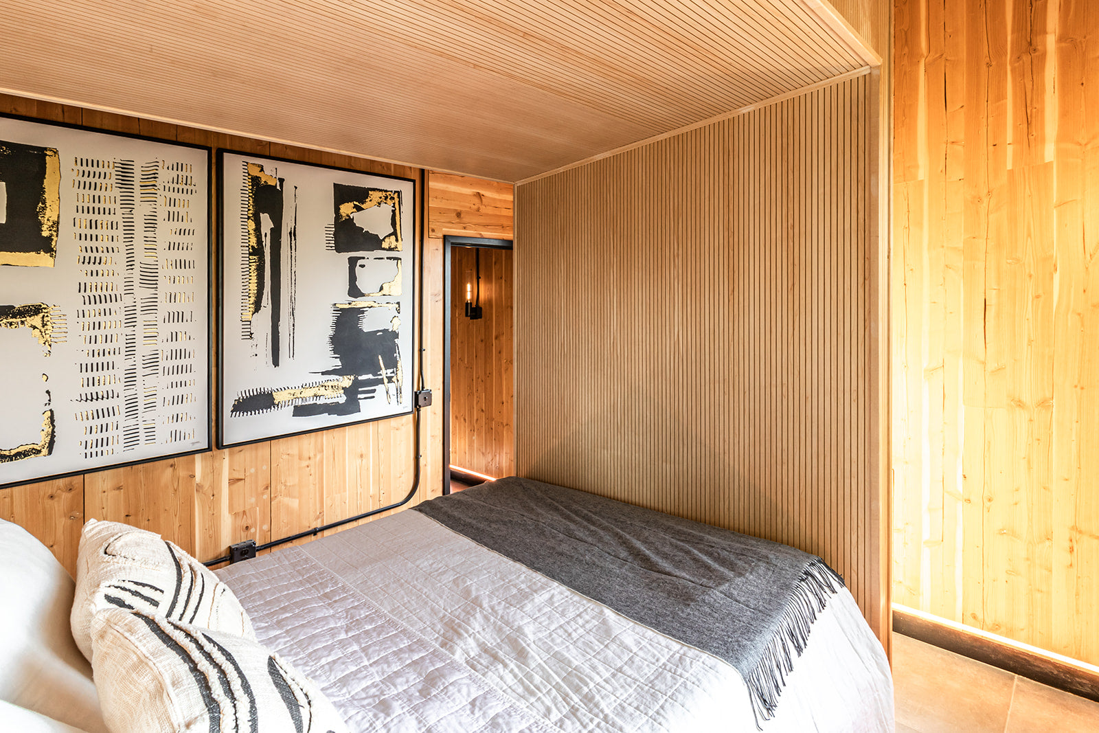 Bedroom with fluted light wood structure going over top of bed, wood paneled walls, and black and white framed artwork with gold accents.