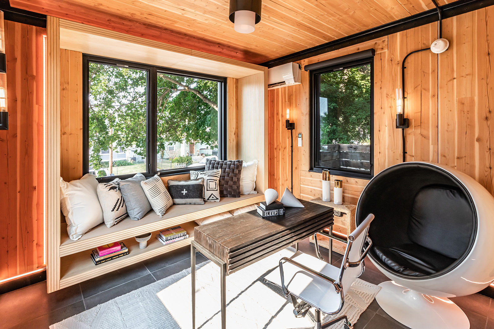 Small home office setup with a bright window and a stylish chair, light wood paneled walls, and elevated wooden bench with neutral pillows and built in storage shelf.