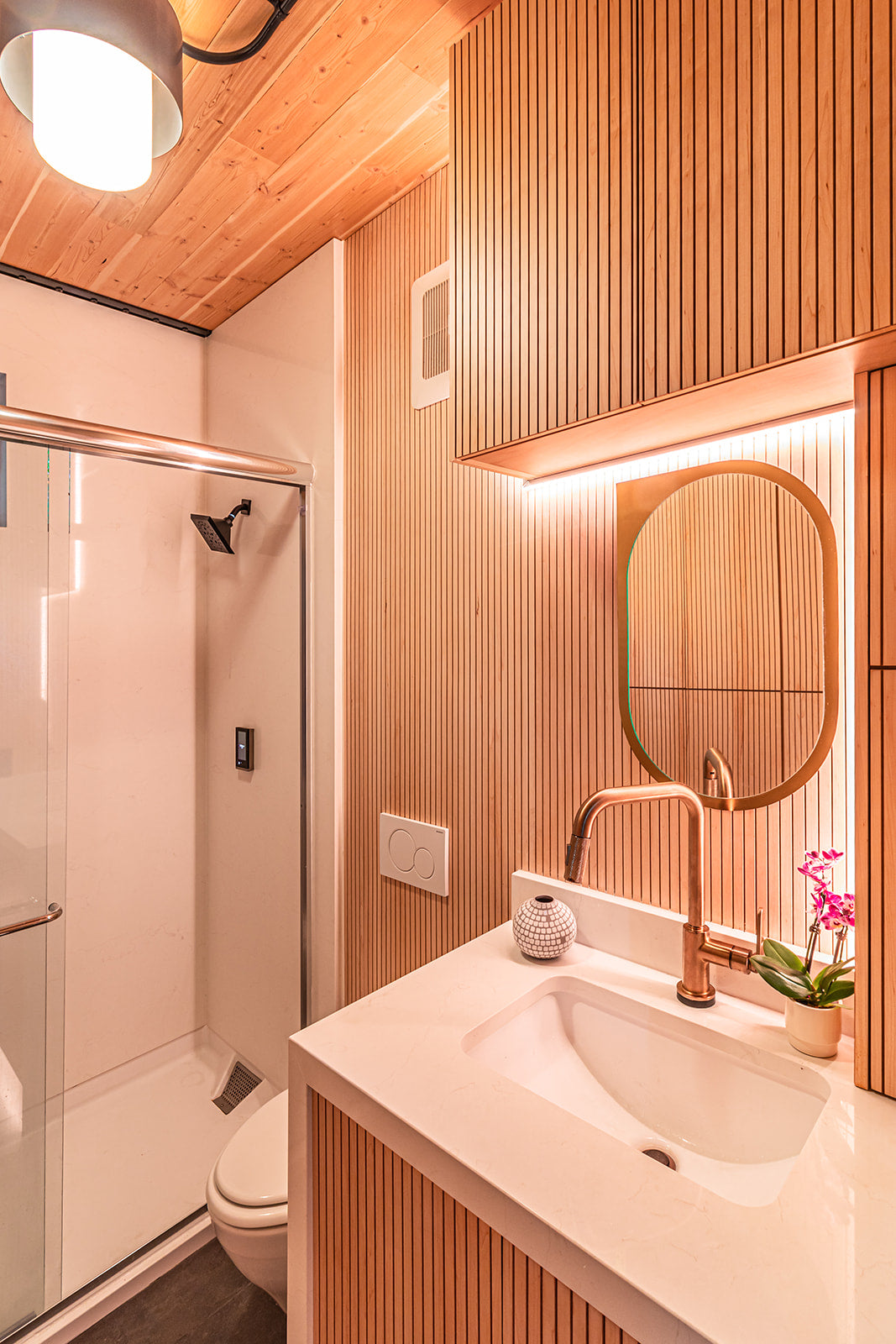 Bathroom with fluted light wood walls, wood paneled ceiling, standing shower with black showerhead, and white vanity sink with gold hardware and mirror.