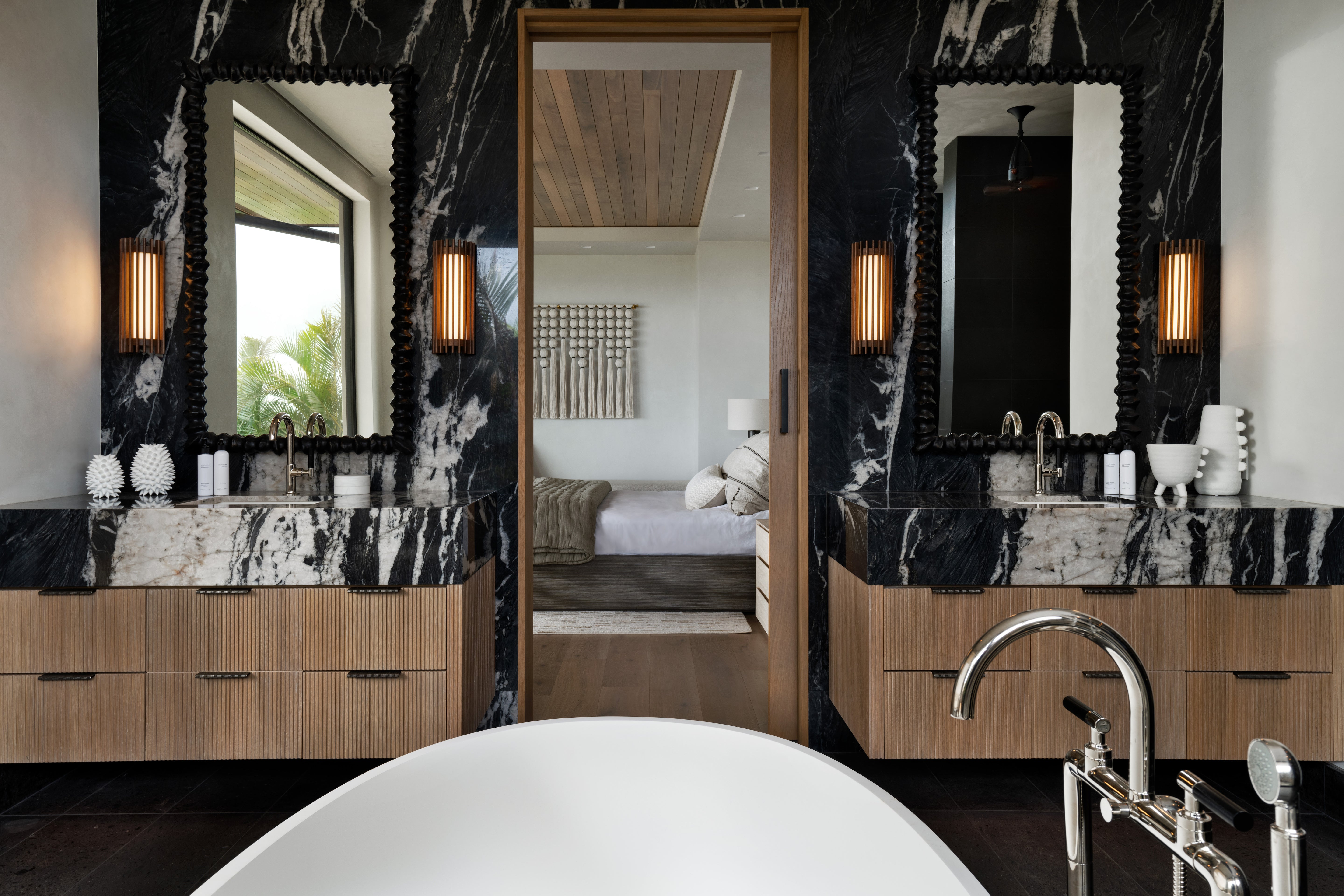 Luxurious bathroom with black and white marble countertops, fluted wood cabinetry, and a freestanding tub, featuring a view into a serene bedroom through a sliding wooden door.