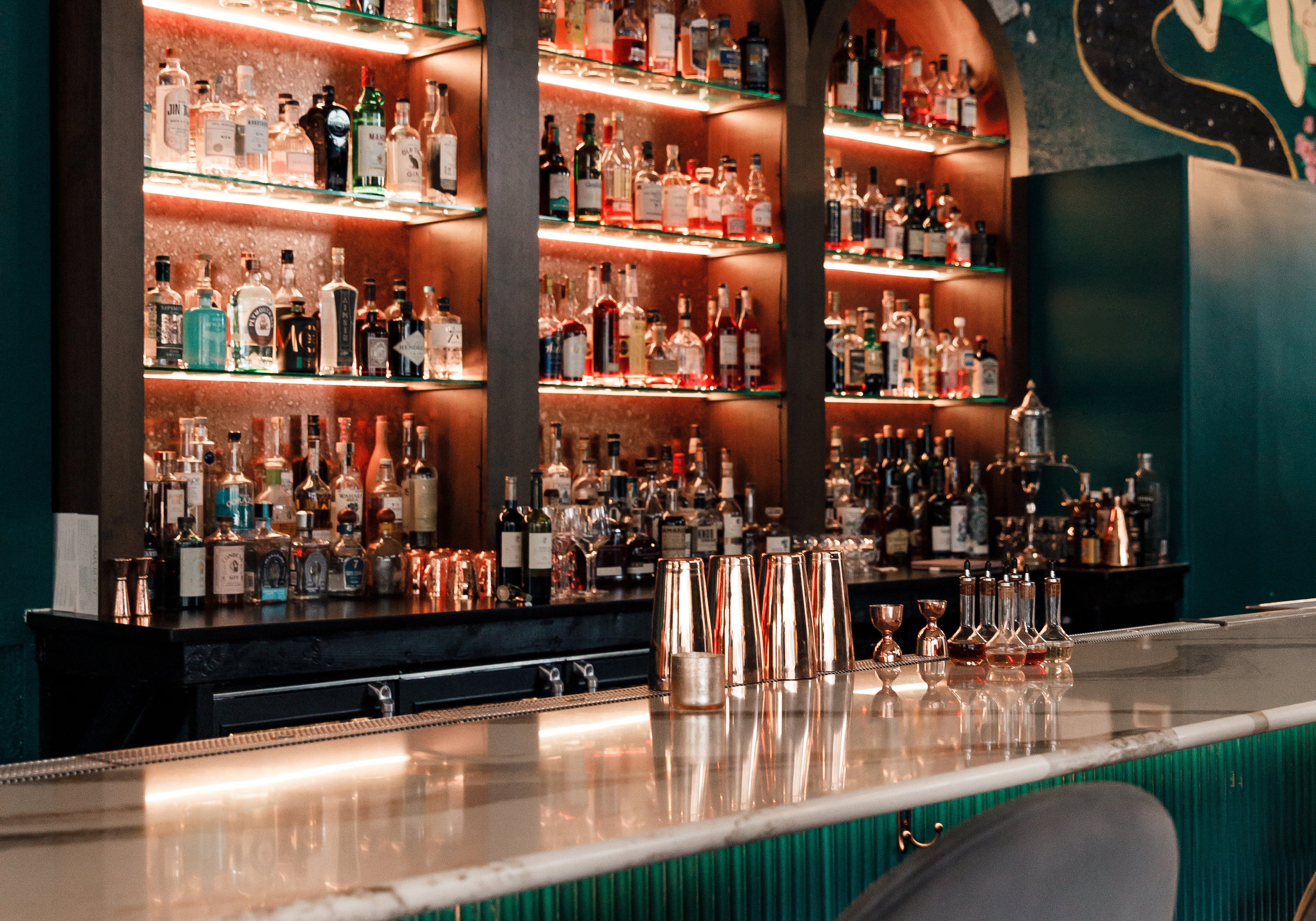 Teal bar with white marble countertops, gray velvet bar stools, dark teal walls mural, and built in glass shelves for liquor.