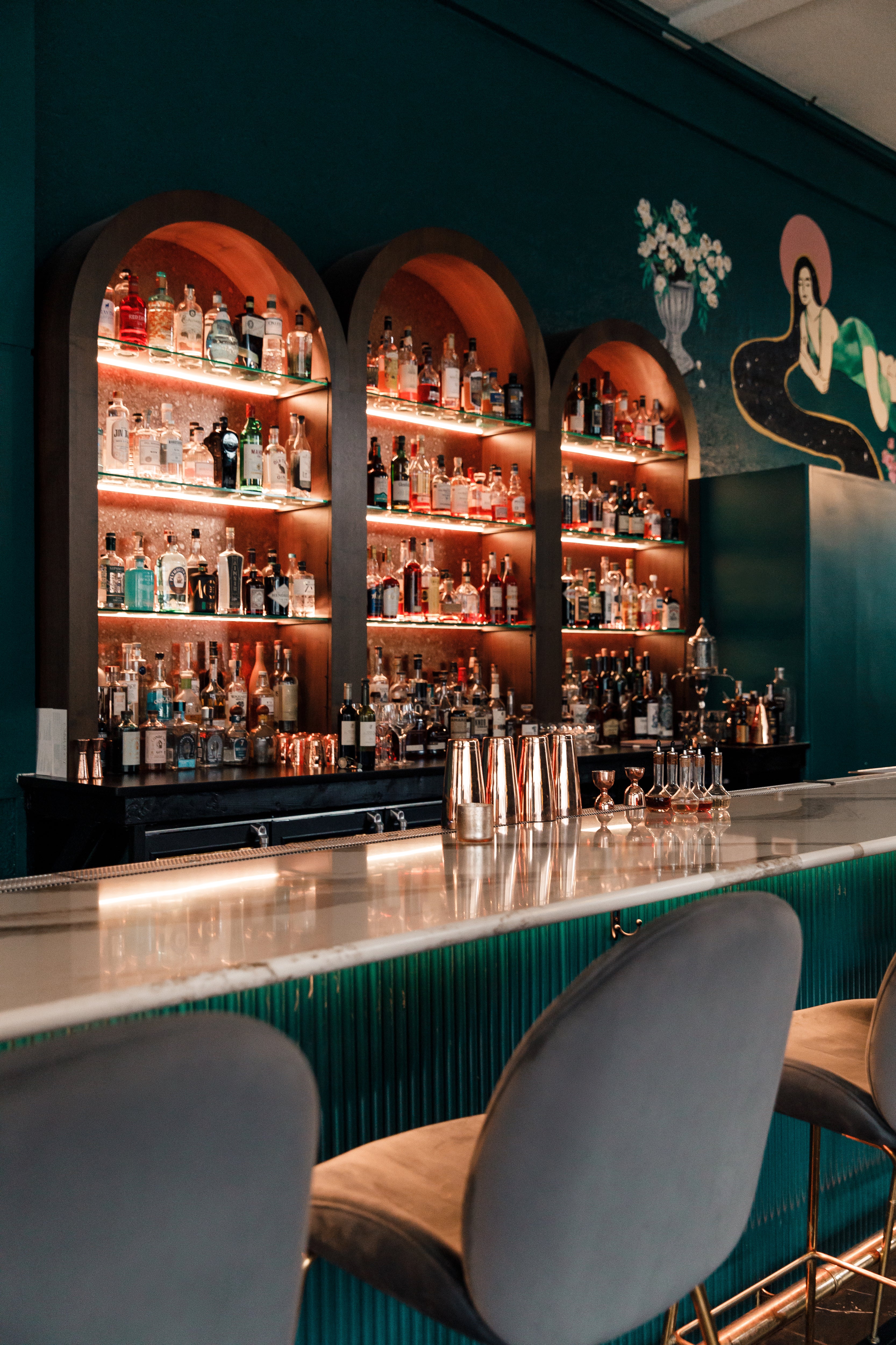 Teal bar with white marble countertops, gray velvet bar stools, dark teal walls mural, and built in glass shelves for liquor.