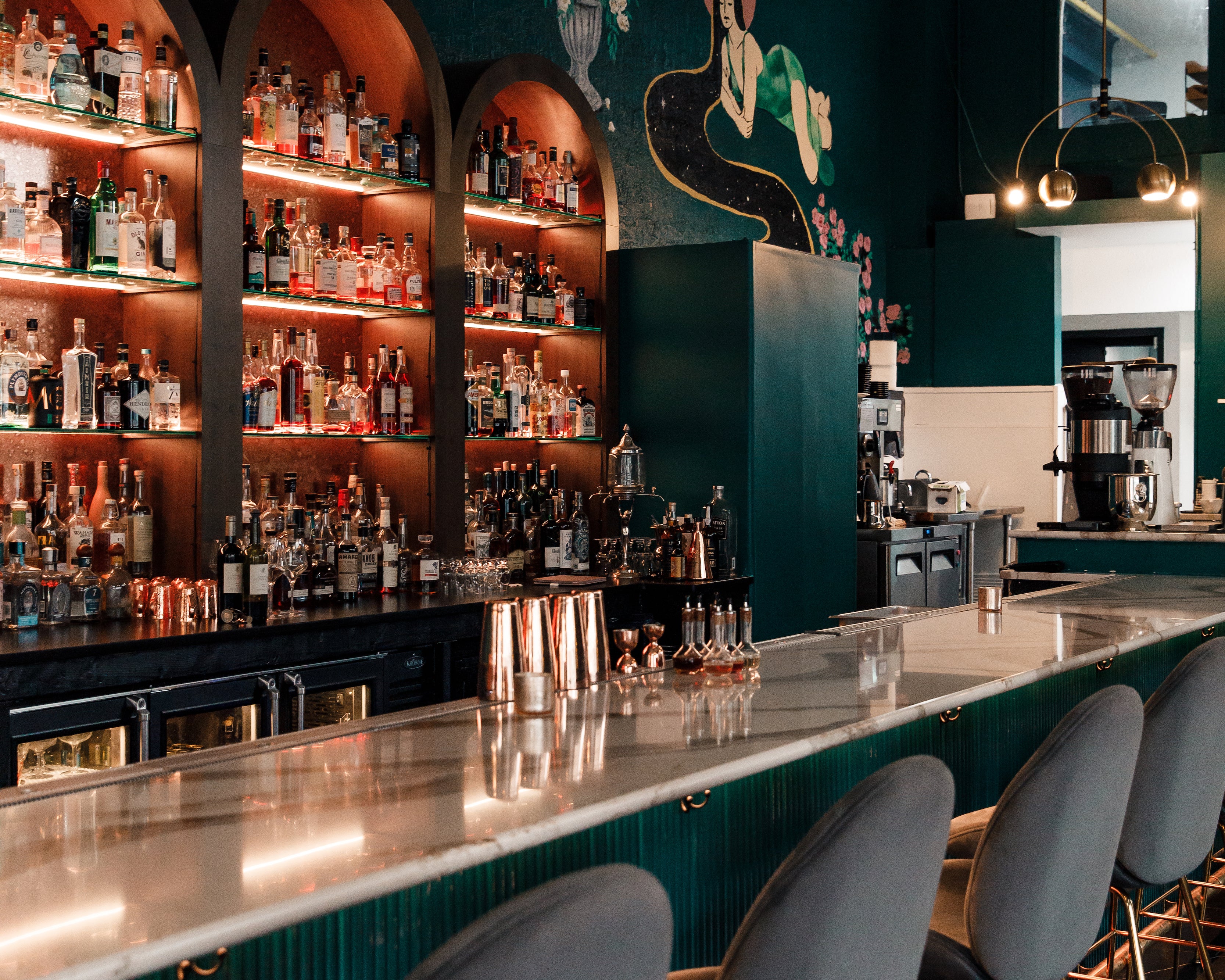 Teal bar with white marble countertops, gray velvet bar stools, dark teal walls with mural, gold pendant lighting, and built in glass shelves for liquor.