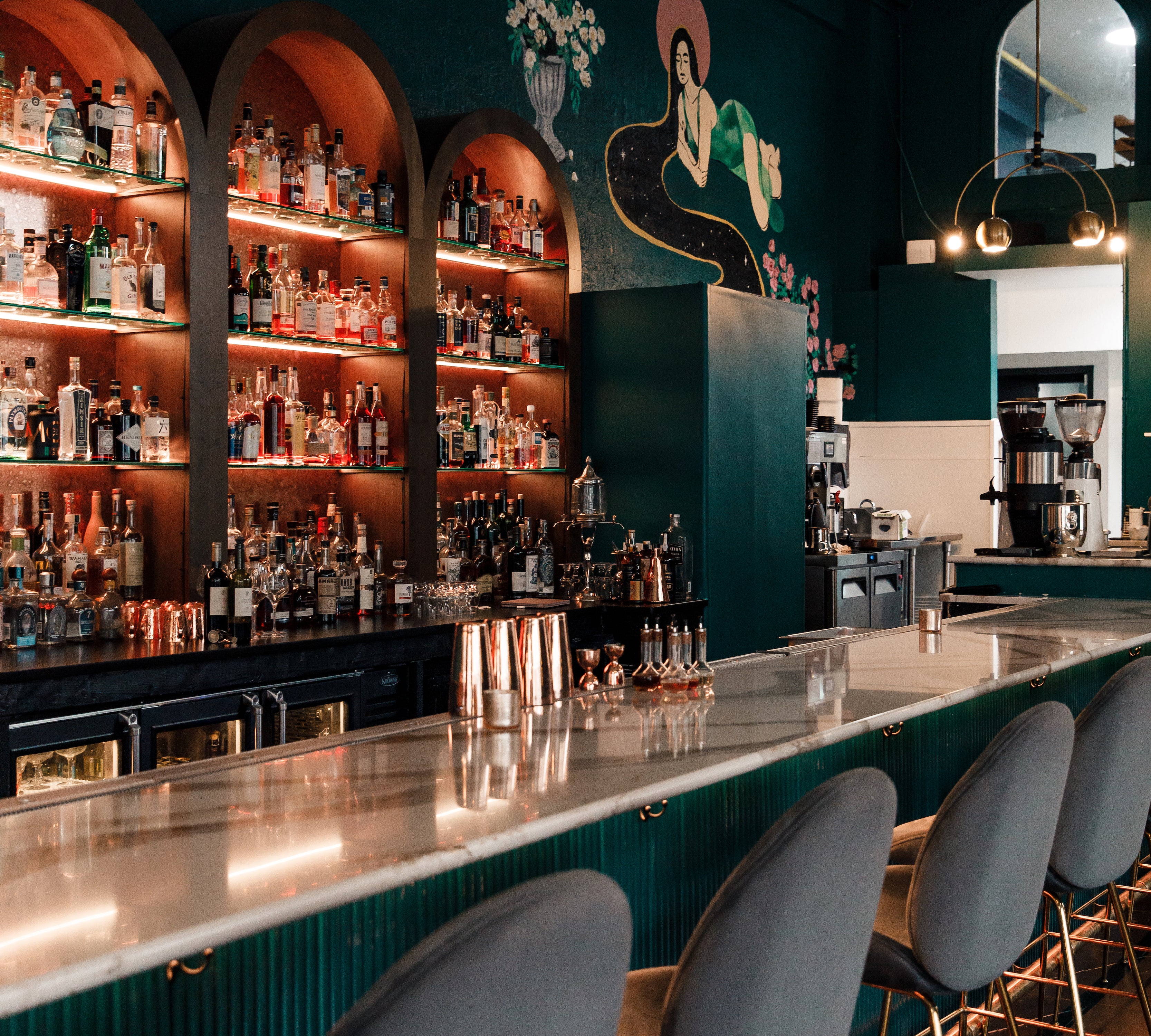 Teal bar with white marble countertops, gray velvet bar stools, dark teal walls with mural, gold pendant lighting, and built in glass shelves for liquor.