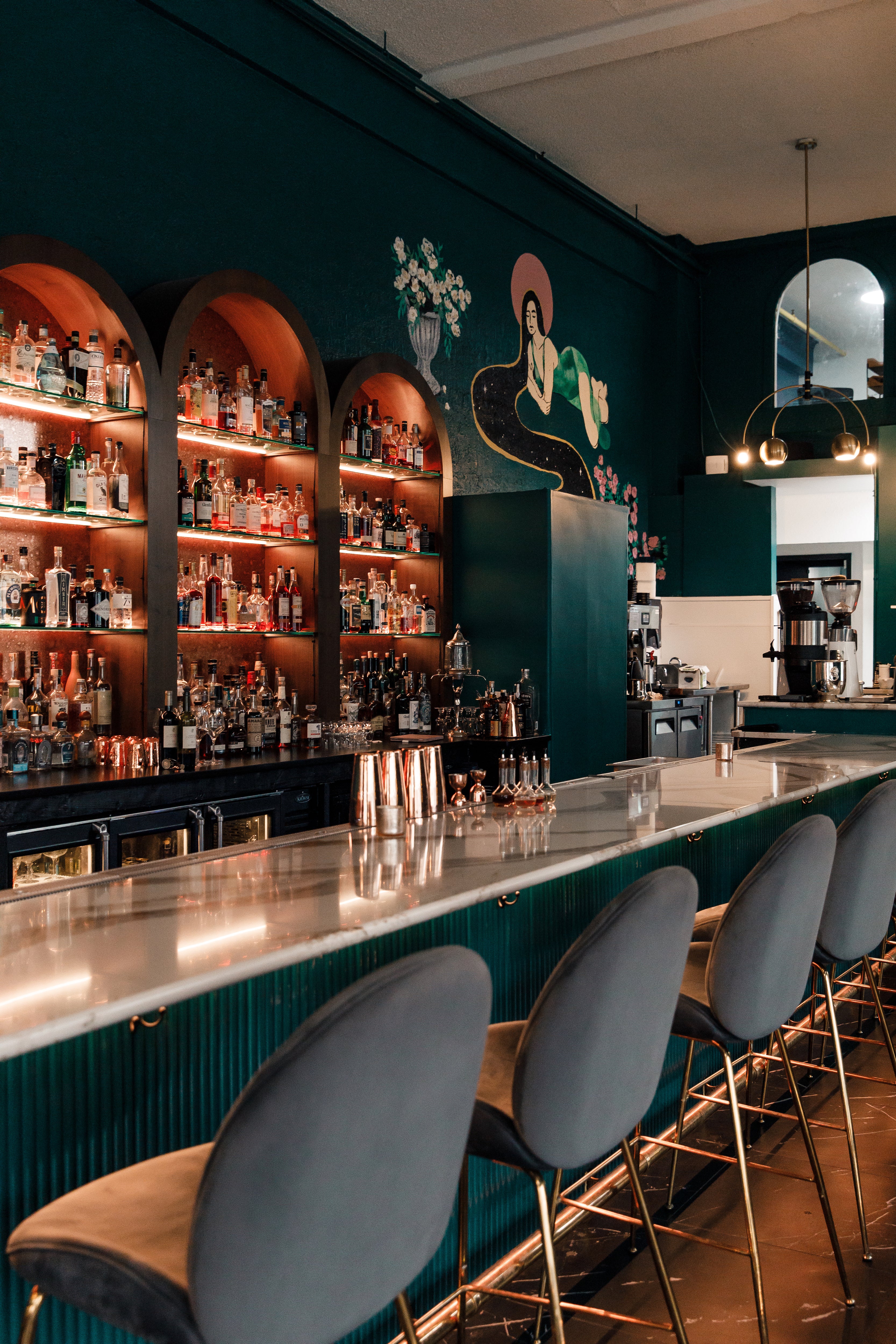 Teal bar with white marble countertops, gray velvet bar stools, dark teal walls with mural, gold pendant lighting, and built in glass shelves for liquor.