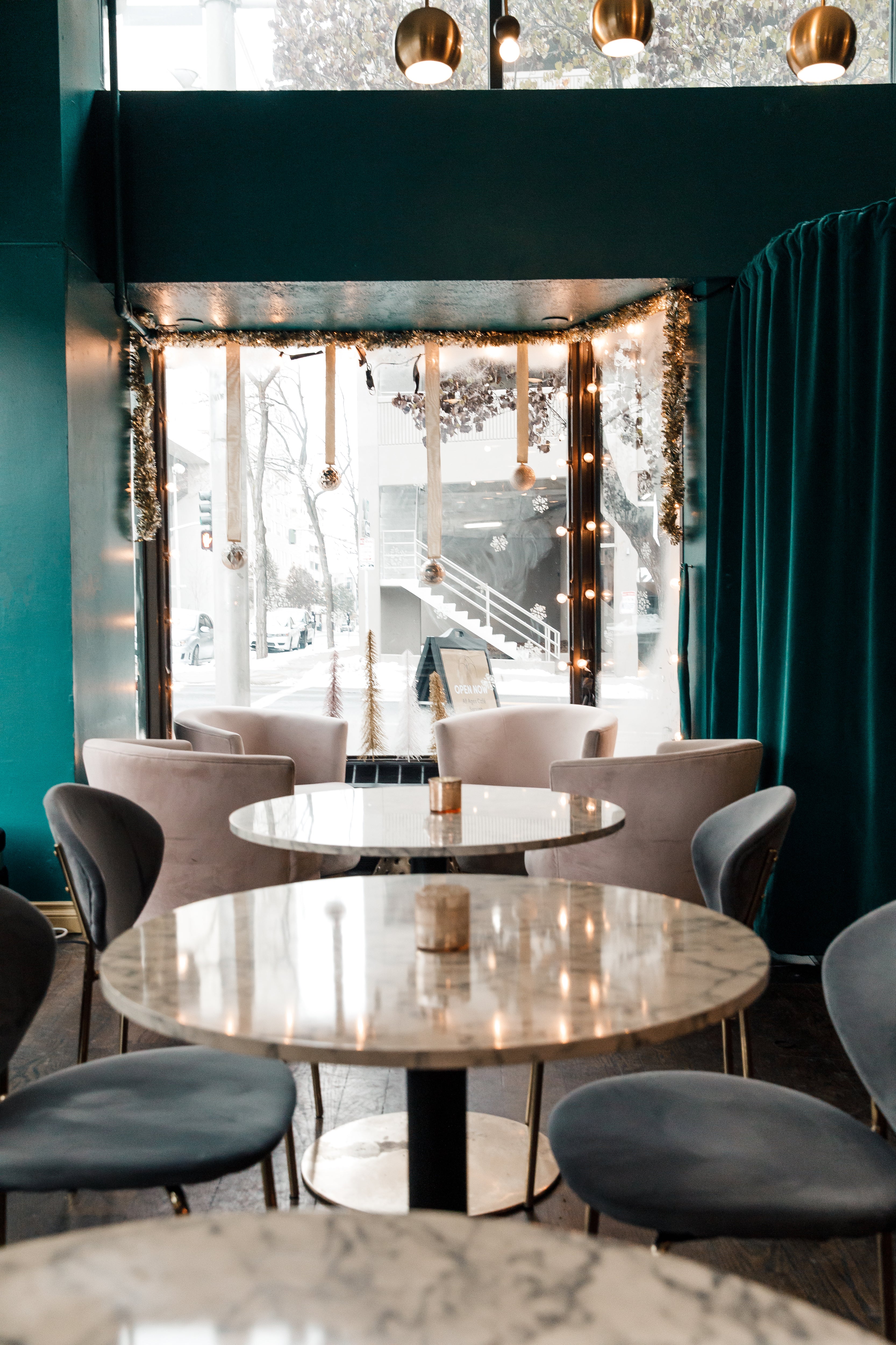 Seating area with small round white marble tables, gray velvet chairs, teal walls, and a bay window with white and silver holiday decor hanging around the window.