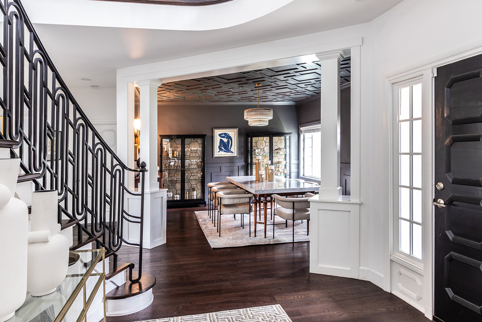 Elegant entryway with a modern black staircase railing, dark wood floors, and a view into a sophisticated dining room featuring a geometric ceiling design, glass cabinets, and a contemporary chandelier.