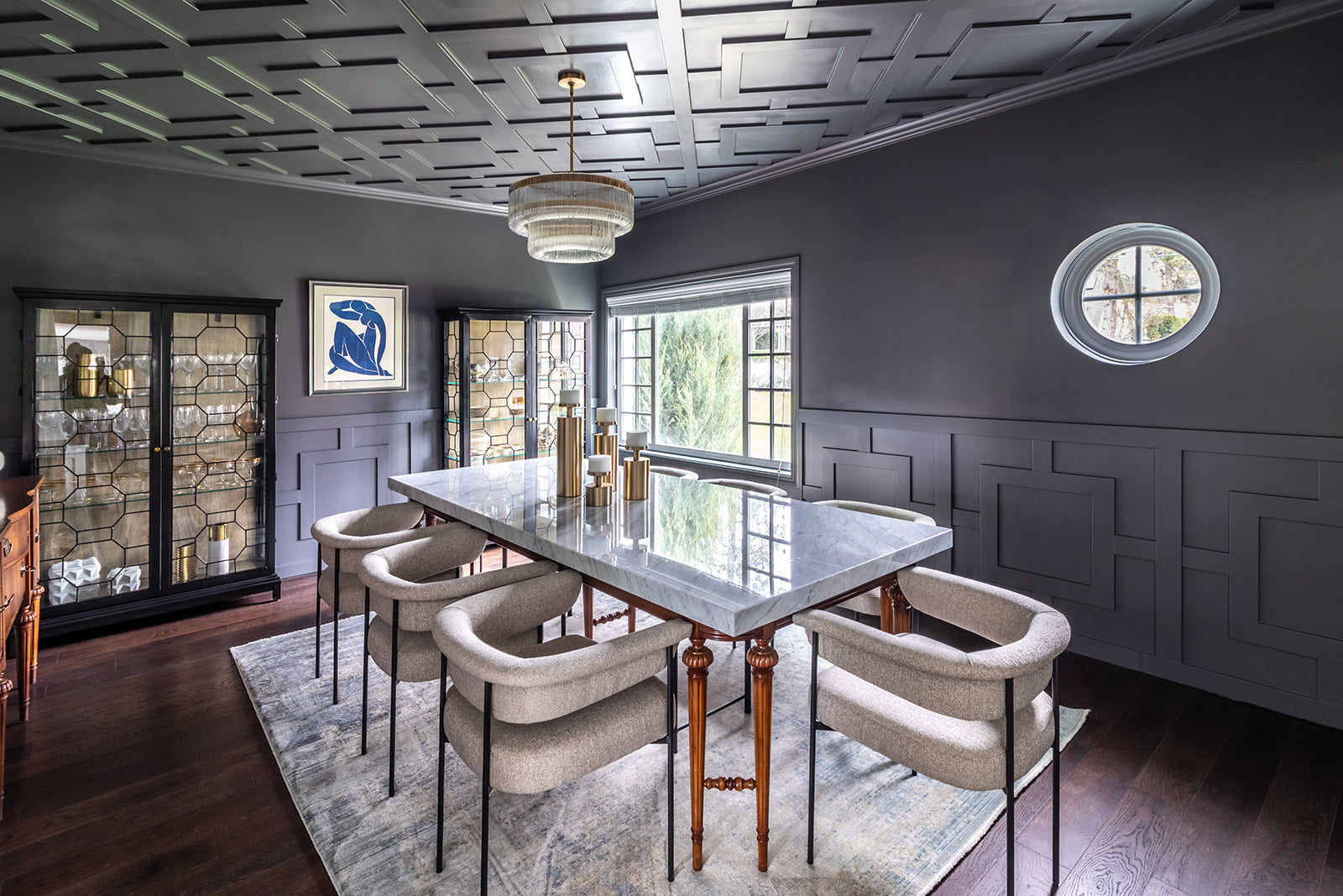 Elegant dining room with a large marble table, modern upholstered chairs, dark paneled walls, and a decorative ceiling design, illuminated by natural light from large windows.