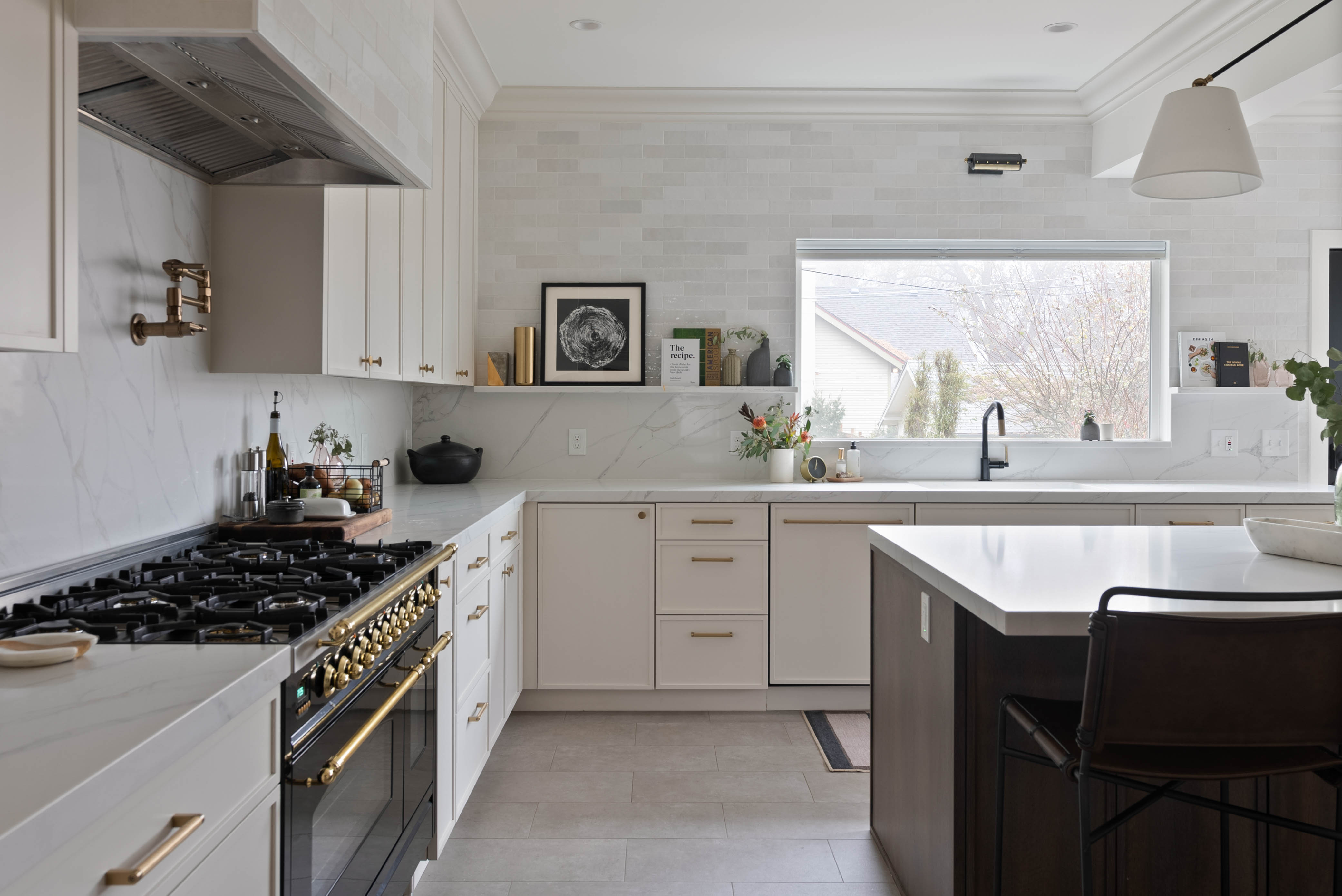 Bright kitchen with white cabinetry, marble countertops, gold hardware, a large gas range, and a central island with seating.