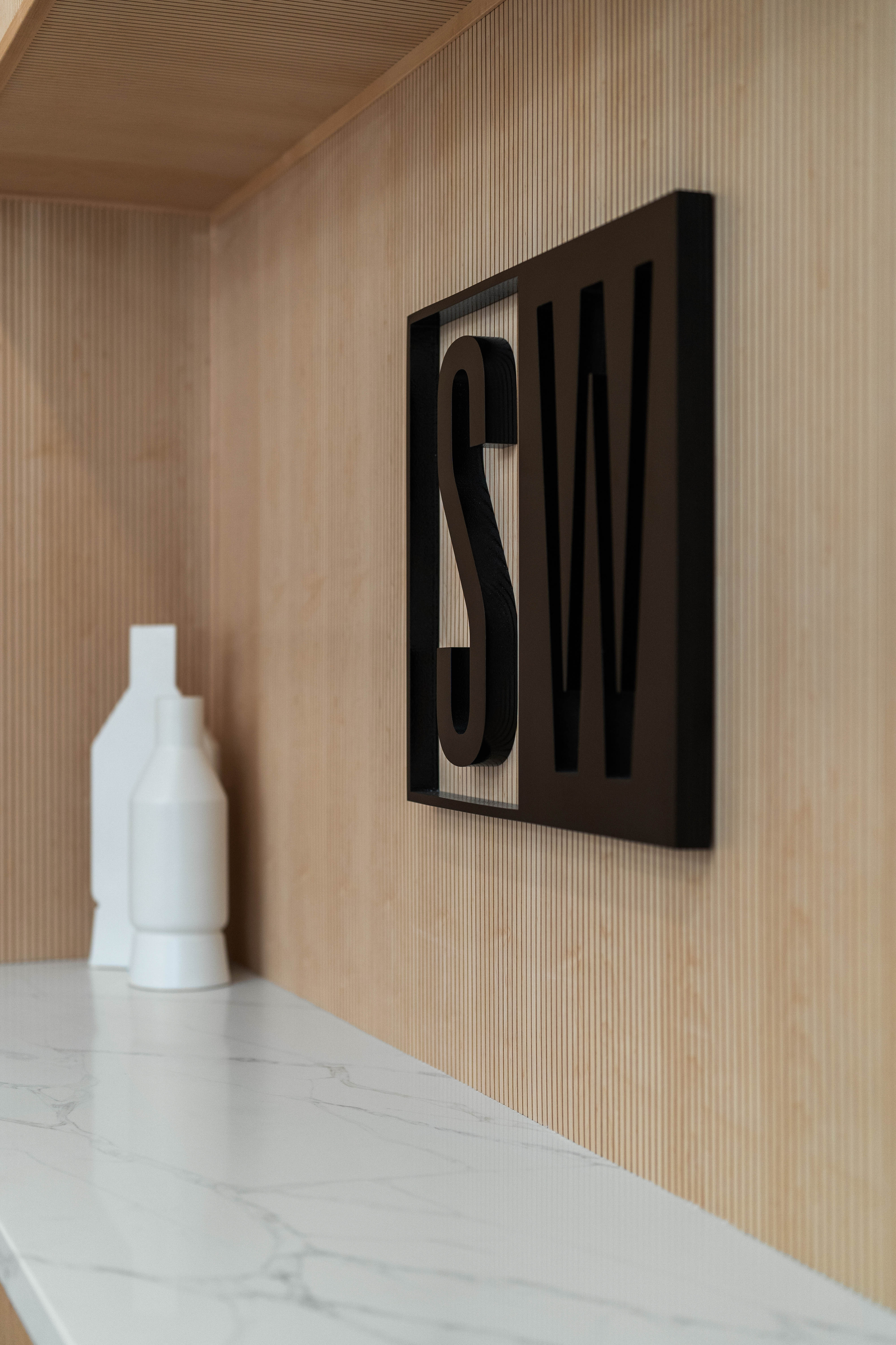 Close-up of black brand logo on fluted light wood wall and white decor on white marble countertop.