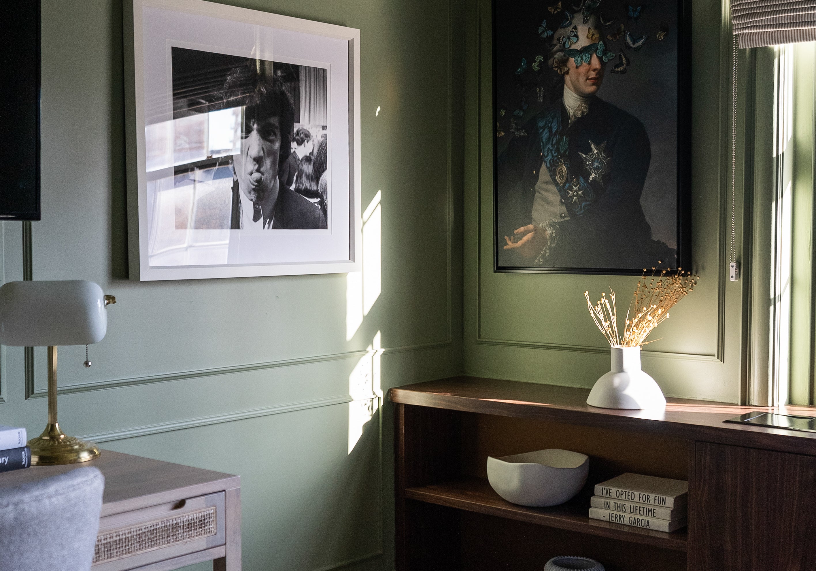 Cozy corner in a bedroom with sage green walls, featuring eclectic artwork, a wooden side table, and soft natural light filtering through the window.