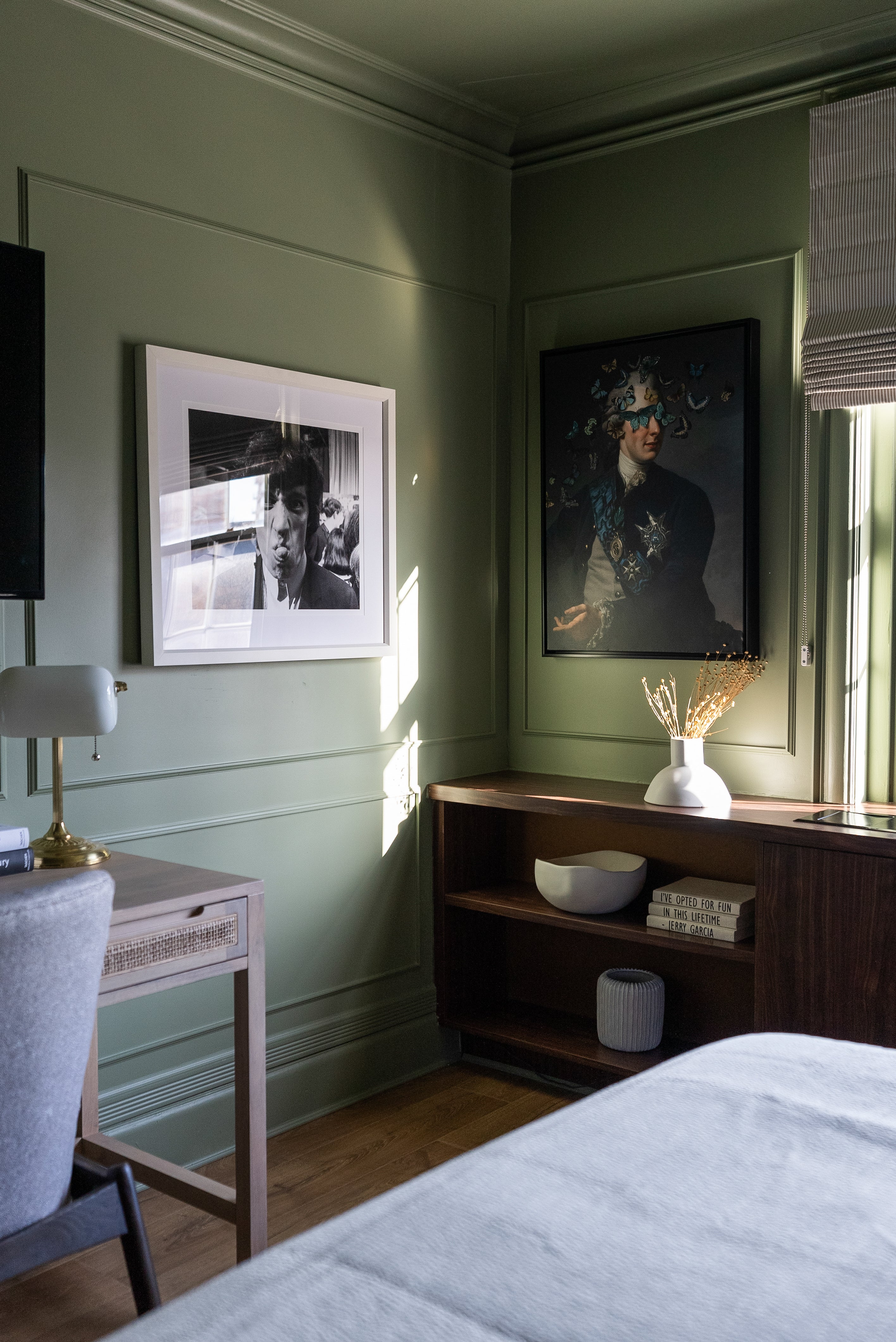 Cozy corner in a bedroom with sage green walls, featuring eclectic artwork, a wooden side table, and soft natural light filtering through the window.