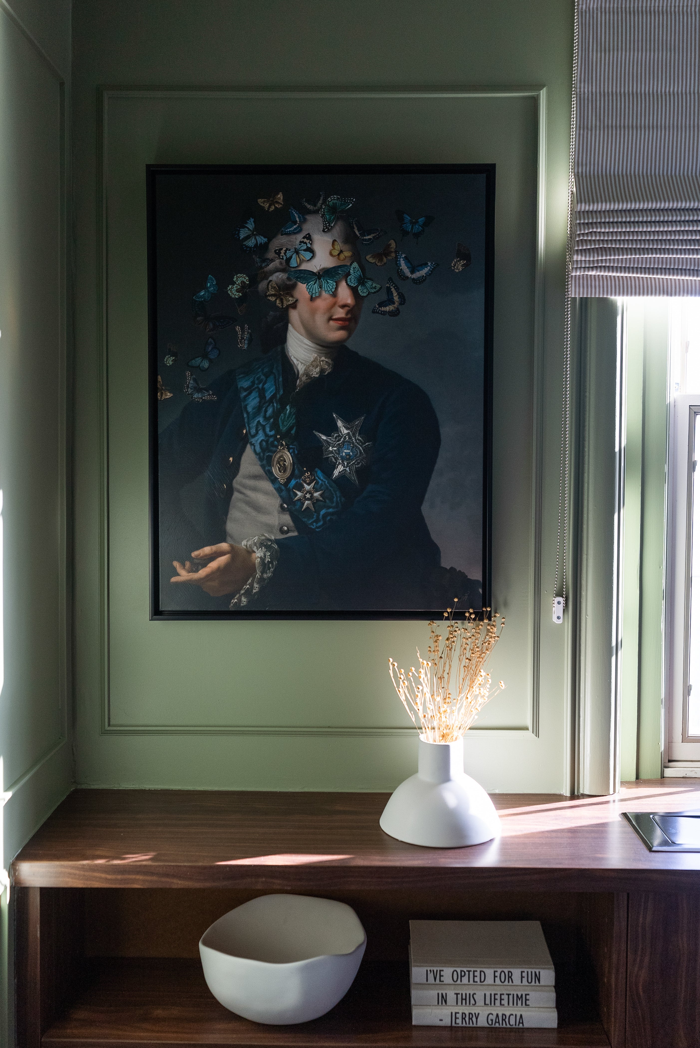 Decorative nook with sage green walls, featuring a whimsical portrait with butterflies, a wooden shelf with minimalistic decor, and natural light filtering through the window.