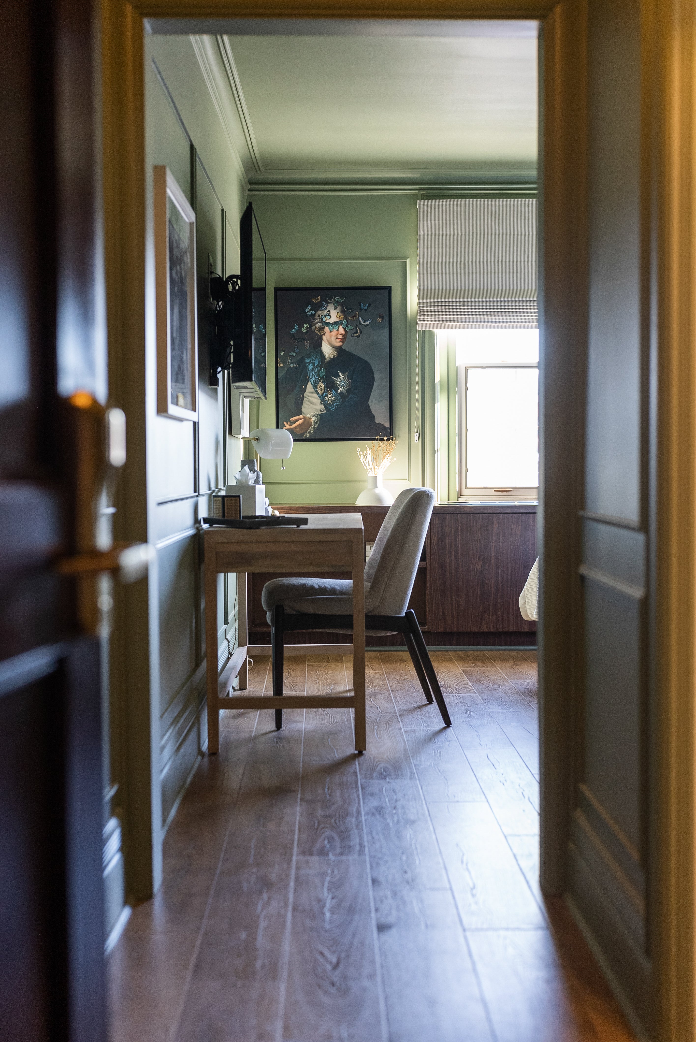 View through a doorway into a cozy room with sage green walls, featuring a small wooden desk, a modern gray chair, and unique artwork on the wall, creating a warm and inviting workspace.