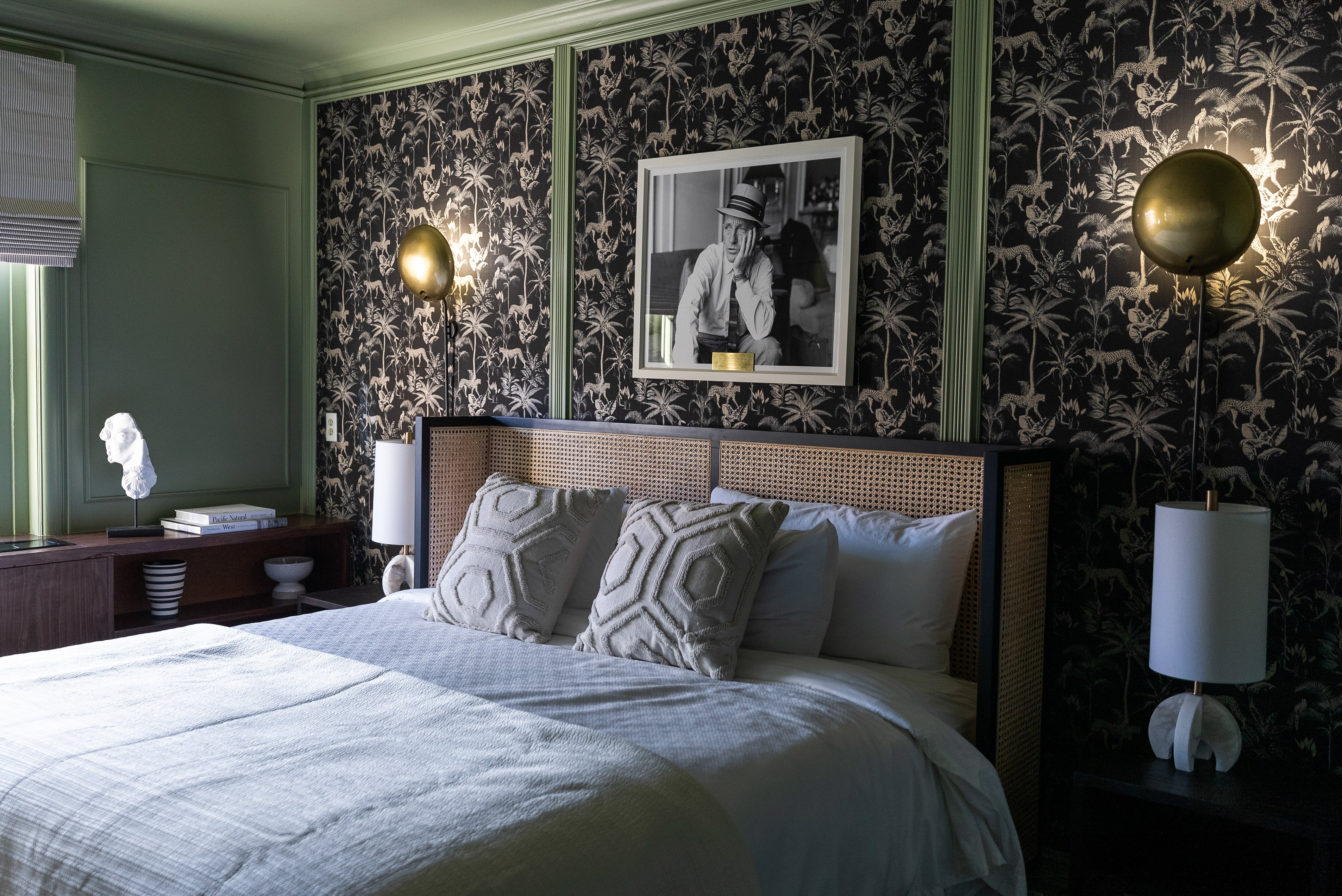 Elegant bedroom with a rattan headboard, geometric pillows, and dark jungle motif wallpaper, accented by gold wall sconces and a black-and-white photograph framed above the bed.