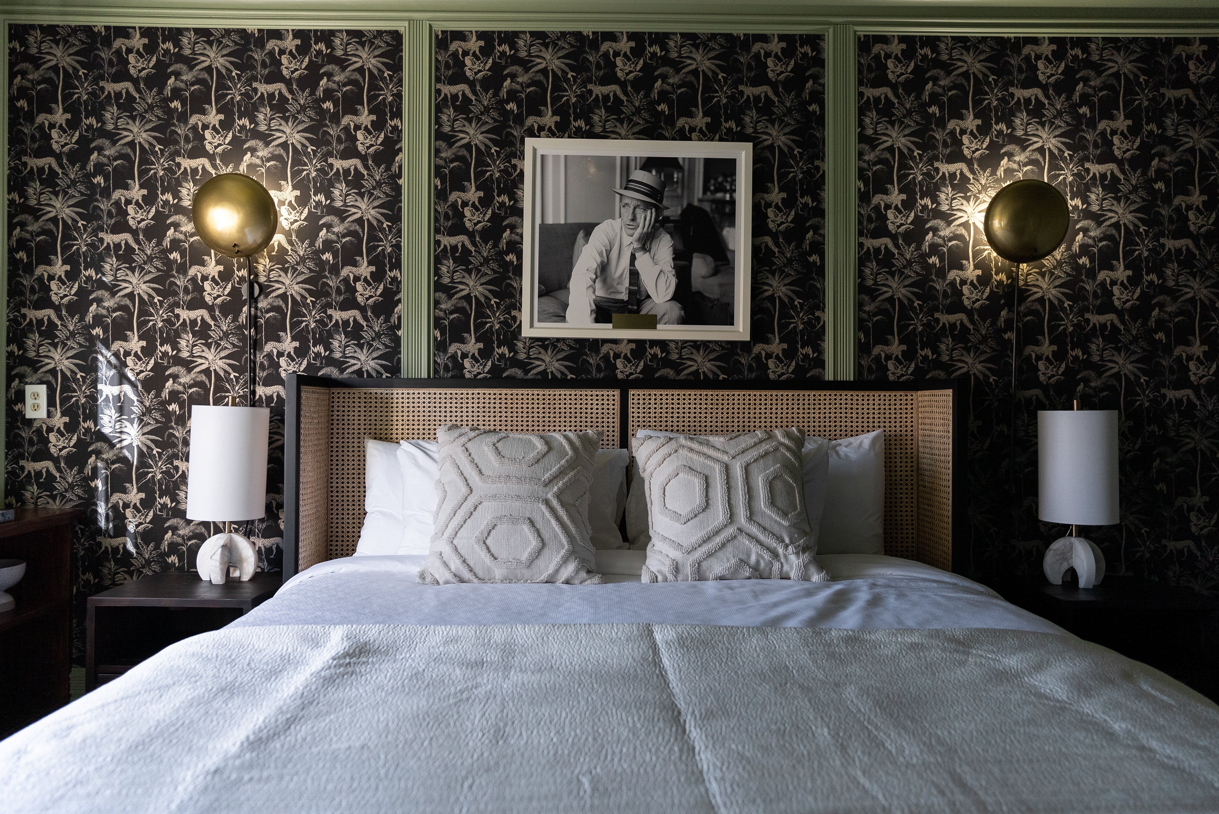 Stylish bedroom with a rattan headboard, geometric pillows, and dramatic black wallpaper featuring a jungle motif, illuminated by gold wall sconces.