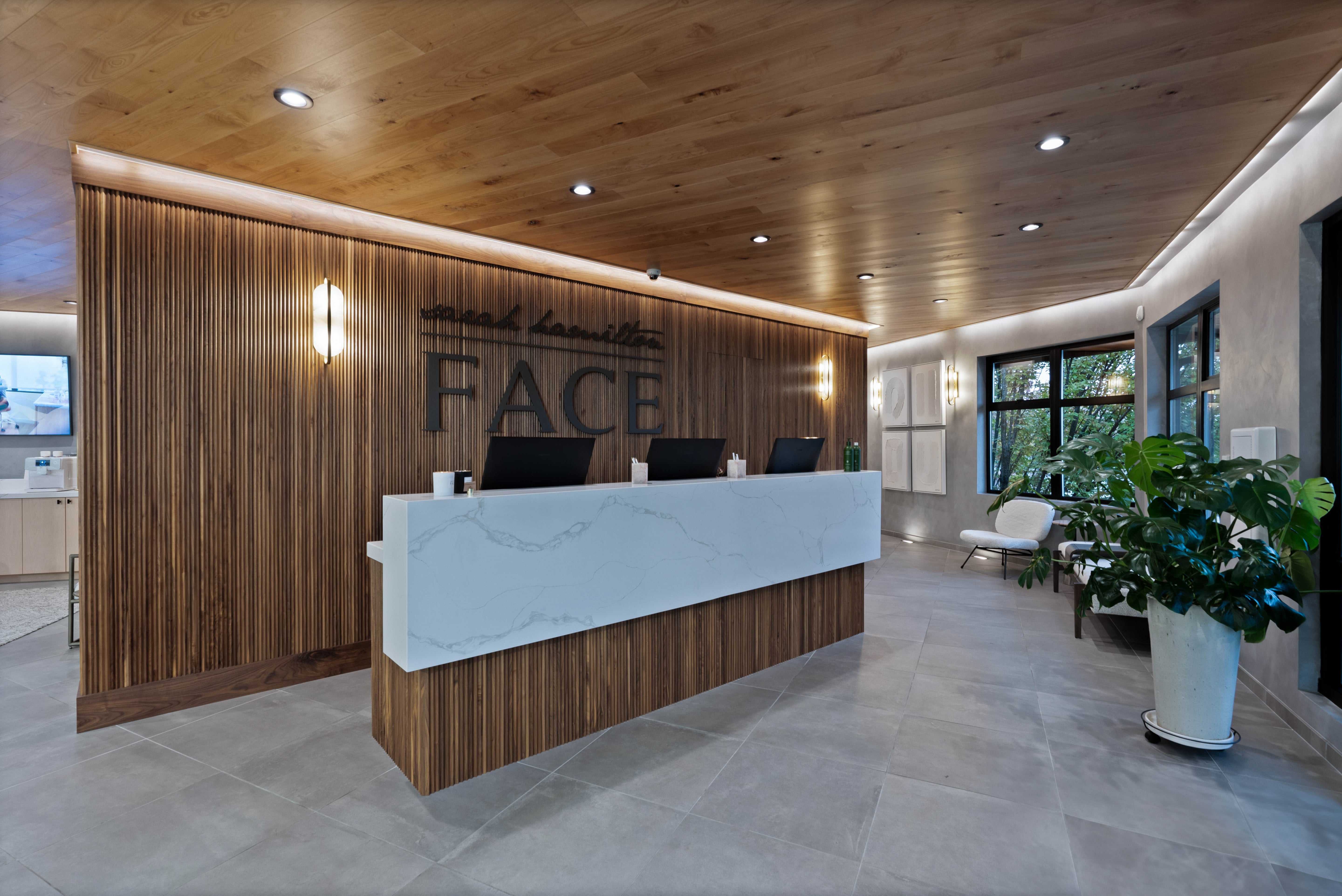 Spacious reception area with a white marble desk, wood slat accent wall with logo on it, warm lighting, and large windows overlooking greenery, creating a serene and professional environment.