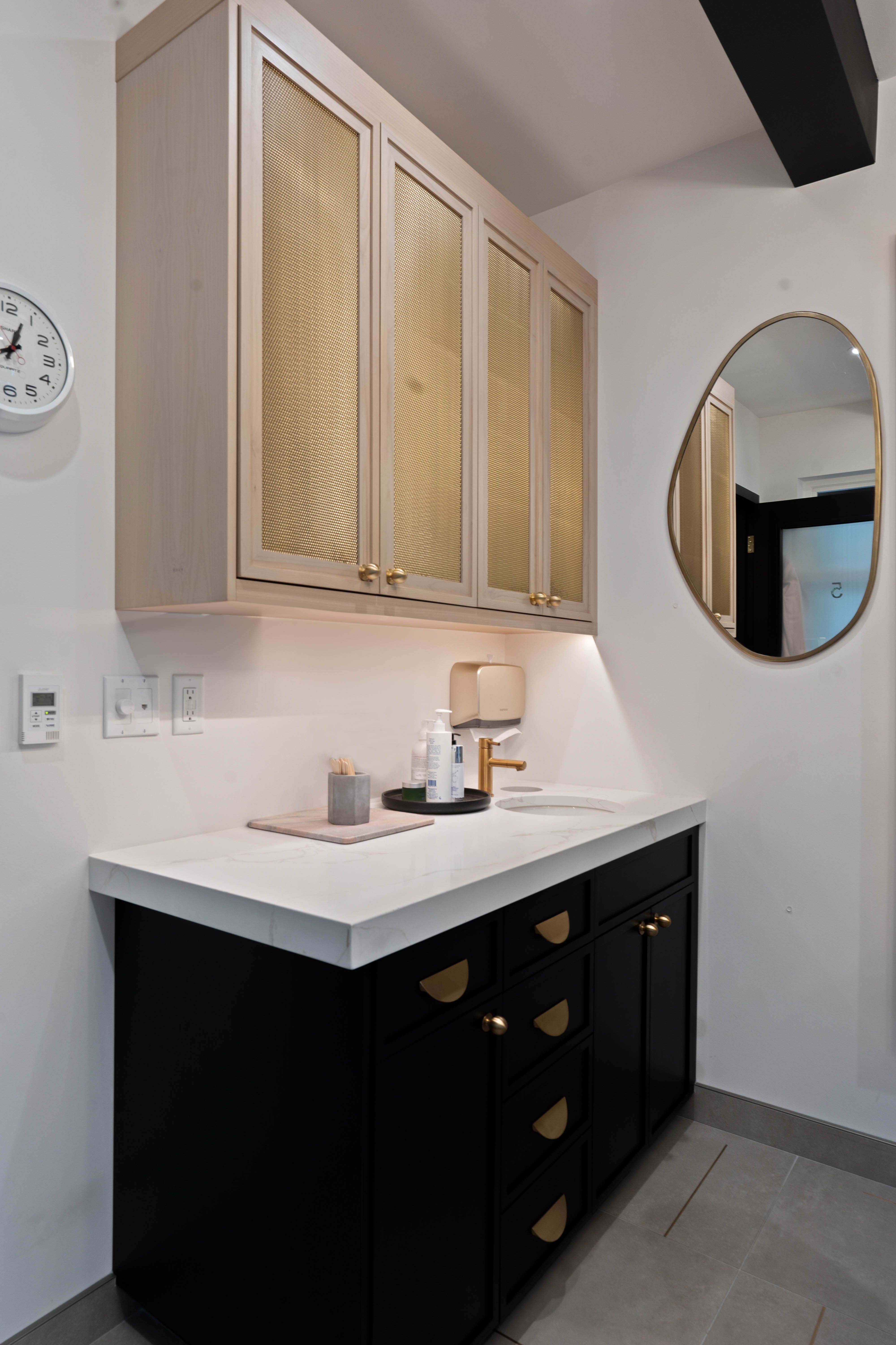 Elegant vanity with black and gold accents, cabinets with gold hardware, topped with a white countertop and complemented by a stylish organic shaped mirror.