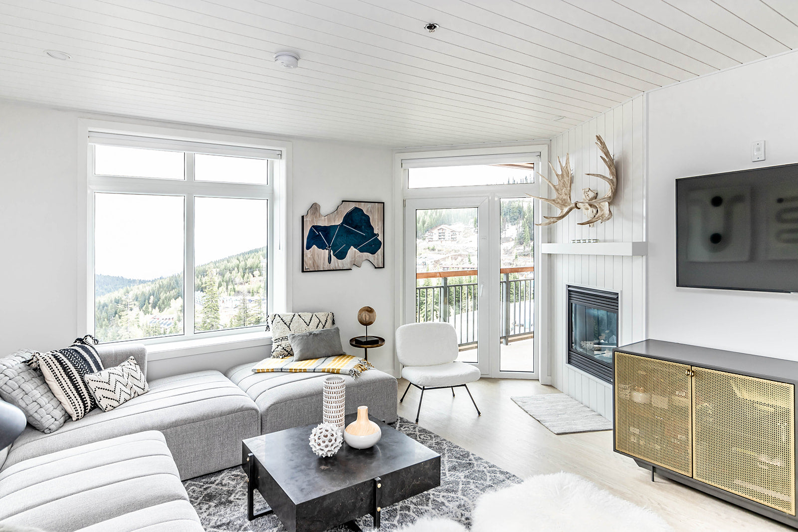 Bright living room with a light gray sectional, fireplace, wall-mounted TV, and large windows offering scenic mountain views, featuring decor and a cozy atmosphere.