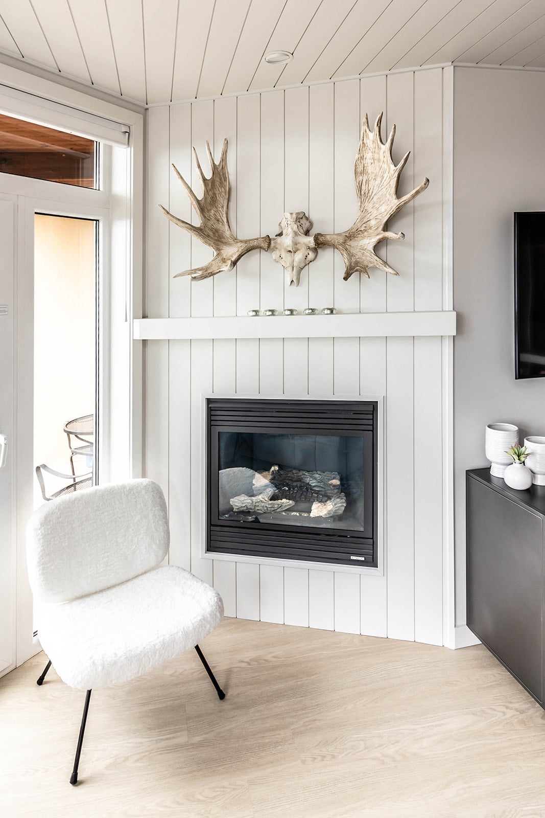 Cozy corner with a modern fireplace, white paneled walls, an antler decor above the mantel, and a plush white chair, creating a warm and inviting atmosphere.