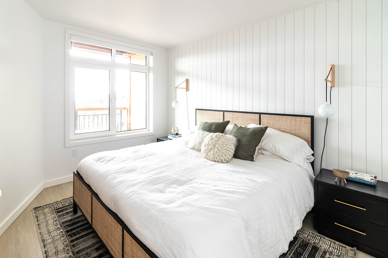 Serene bedroom with a white paneled accent wall, a large window, a bed with a rattan headboard, and cozy green and cream pillows, creating a calm and inviting space.