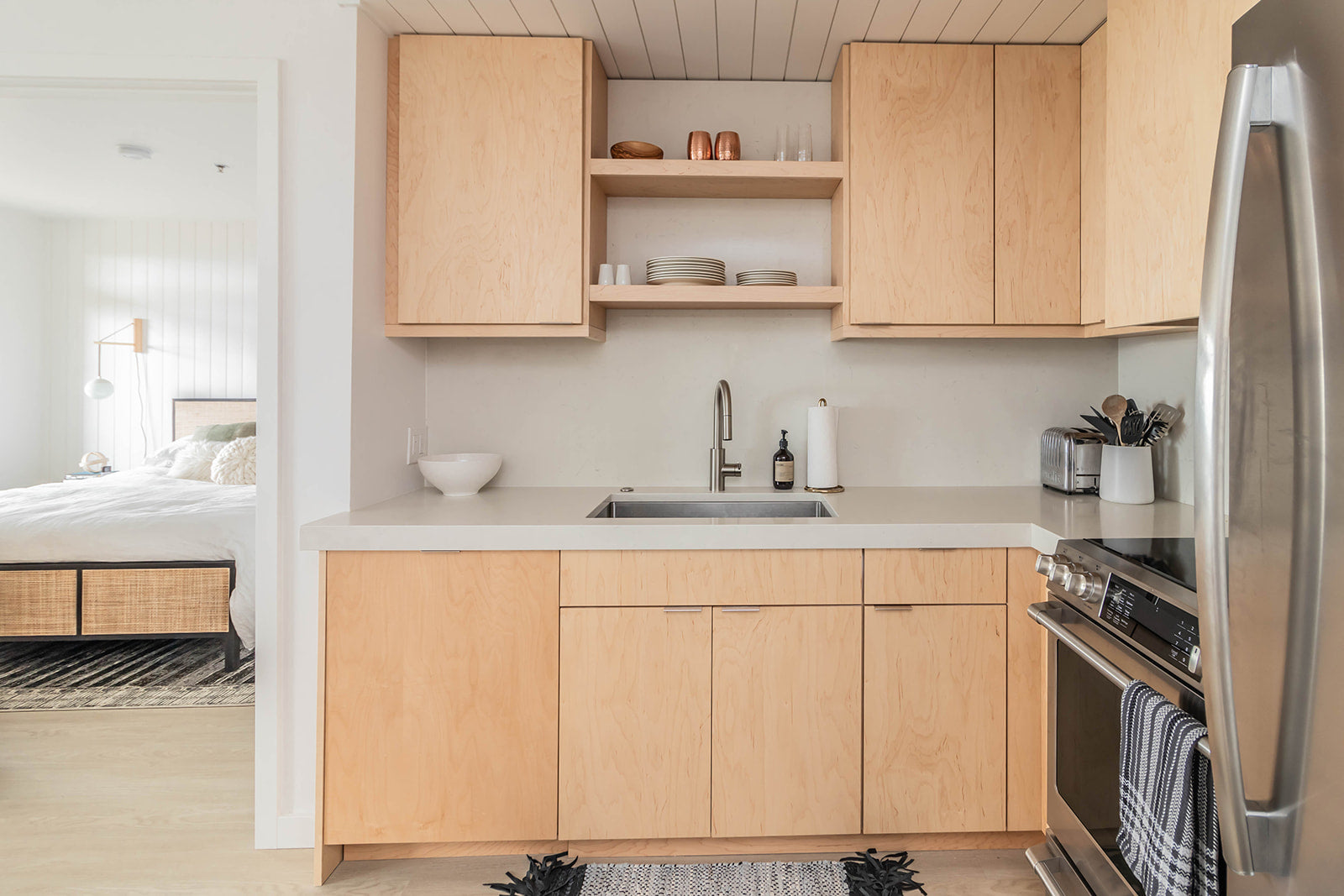 Kitchen with light wood cabinets, open shelving, and stainless steel appliances, adjacent to a cozy bedroom with a minimalist design.