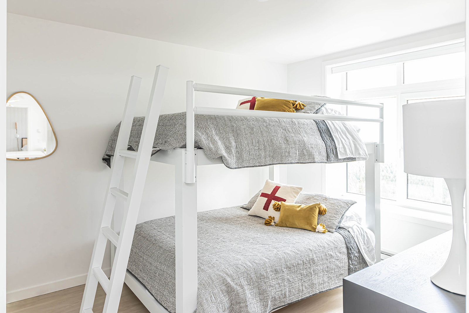 Bright bedroom with white bunk beds, cozy gray bedding, and decorative pillows, featuring a large window that brings in natural light and a minimalist design.