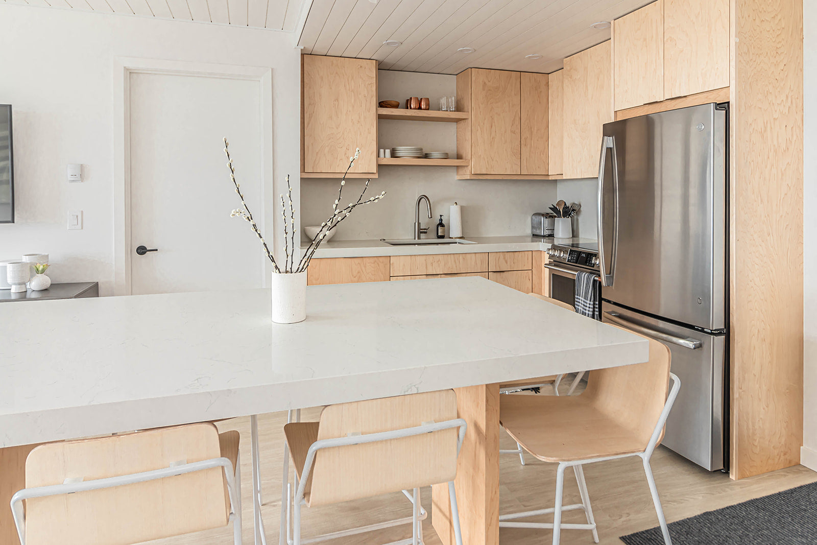 Kitchen with light wood cabinetry, a large white island, stainless steel appliances, and minimalist decor, featuring a white vase with branches as a centerpiece.