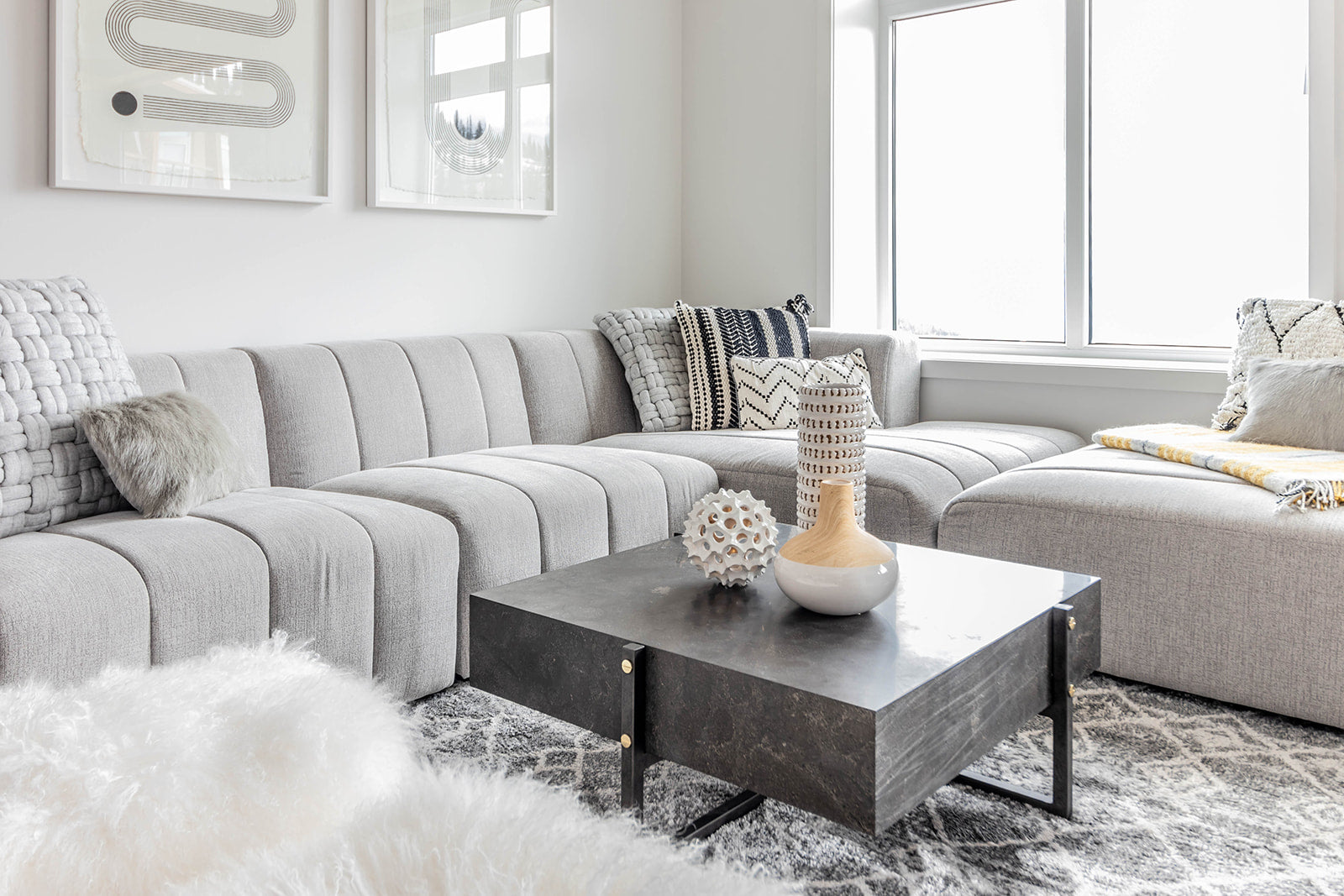 Cozy living room with a light gray sectional sofa, patterned throw pillows, and a black coffee table adorned with decorative vases, set against large windows that bring in natural light.