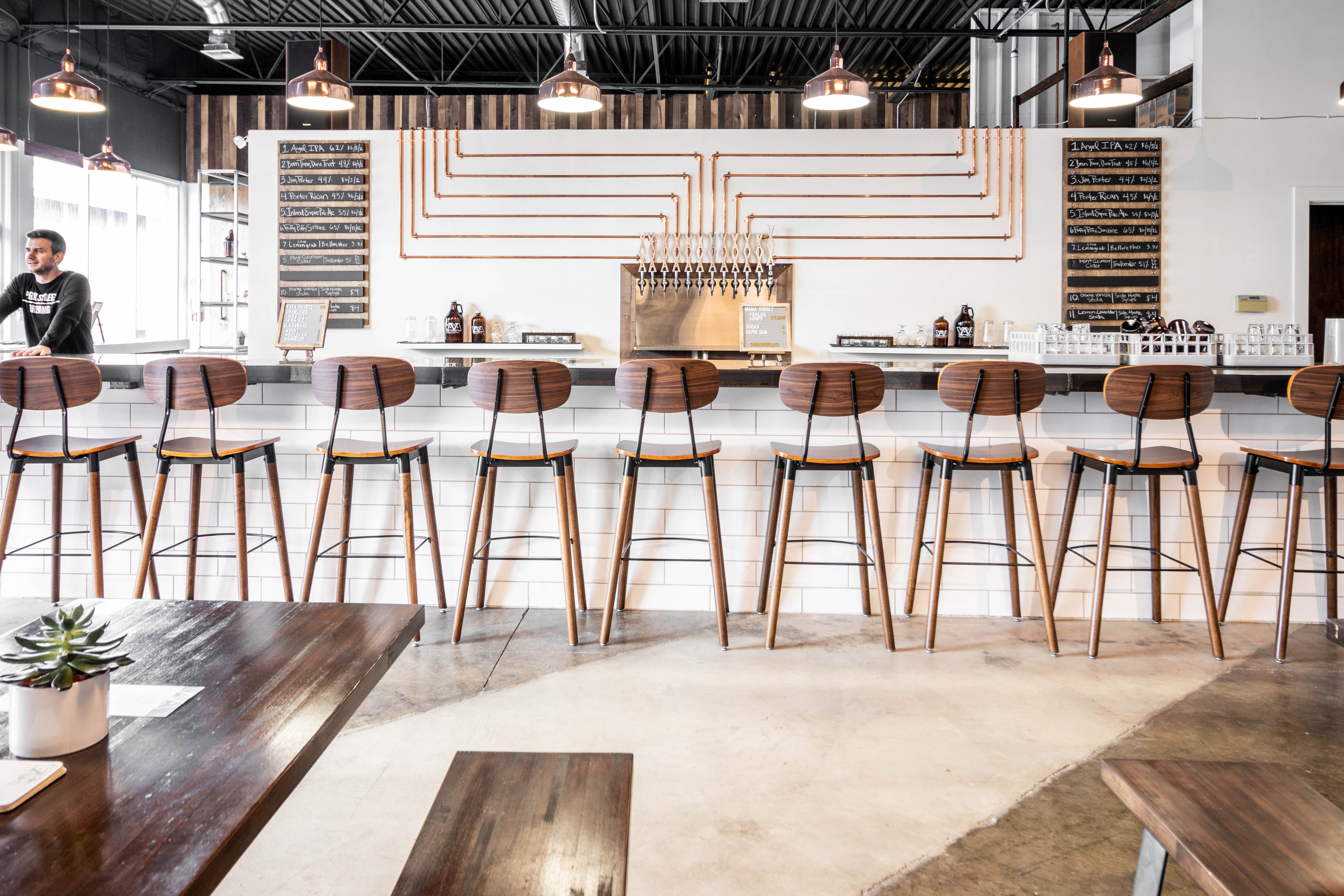 Long bar counter with white subway tiles, wooden barstools, and white walls with chalkboard drink signs and copper pipes on walls leading to draft.