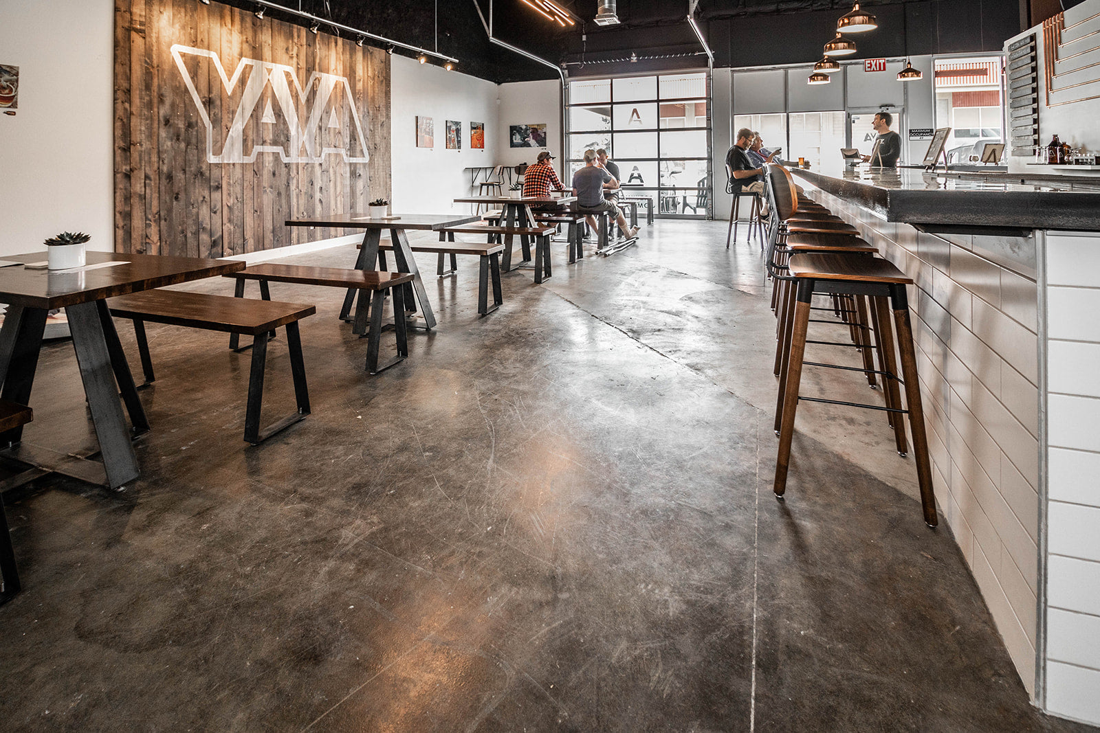 Bar with white subway tile, wooden bar stools, and black countertops. Seating area with wooden tables and benches, white walls with small artwork, and large accent section on wall with wood paneling and brand logo.